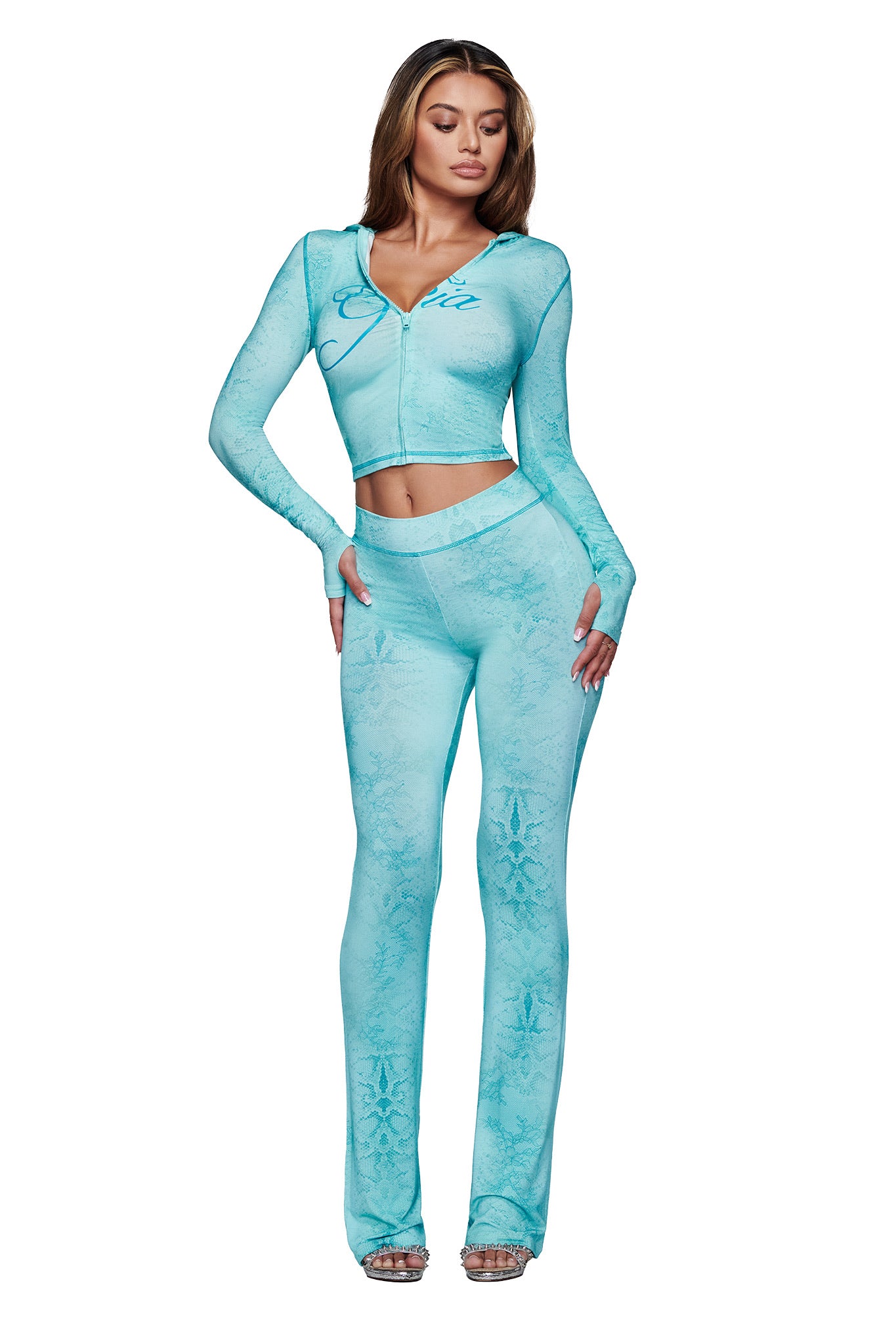 A woman models the I.AM.GIA BLARE TRACKPANT - TEAL SERPENTINE, a fitted two-piece with long sleeves and flared legs, styled with silver heels against a white background.