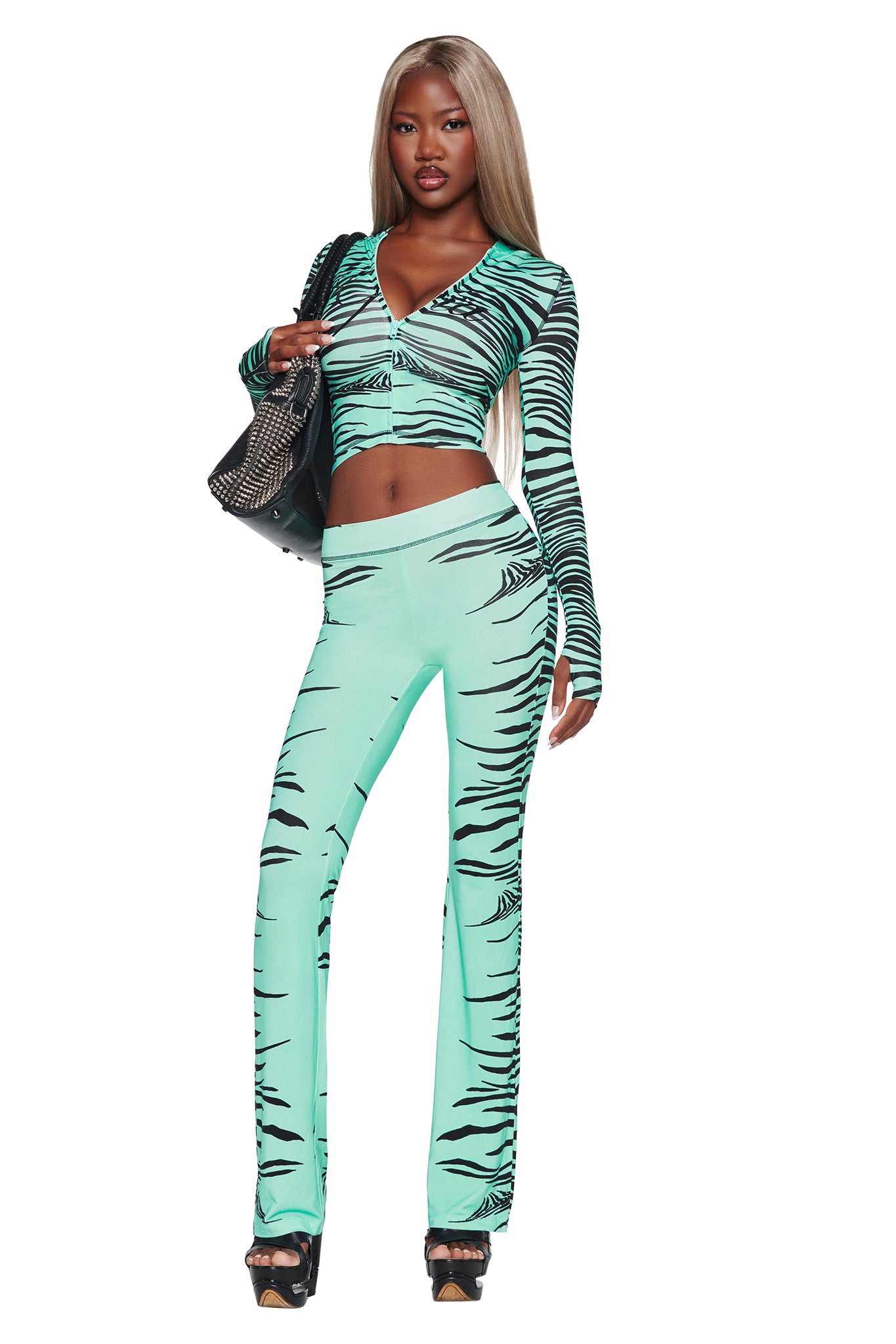 A woman stands confidently against a white background, wearing the I.AM.GIA BLARE TRACKPANT in Aqua Fine Line Zebra—a turquoise, zebra print matching set with long sleeves and flared pants. She accessorizes with a black handbag and black high heels.