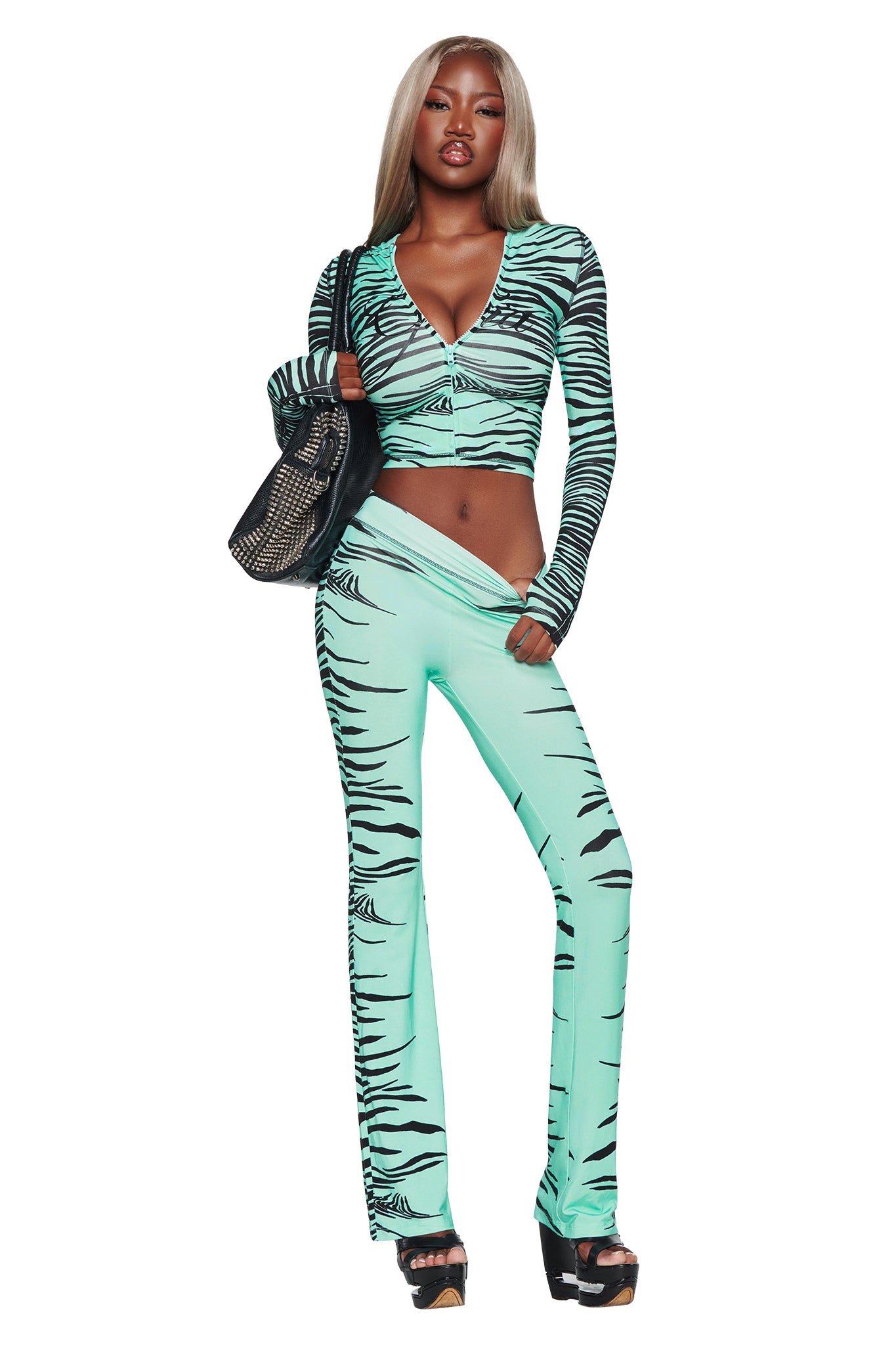 A woman dressed in the I.AM.GIA BLARE TRACKPANT in Aqua Fine Line Zebra, holding a black purse and wearing black platform heels, stands against a white background.