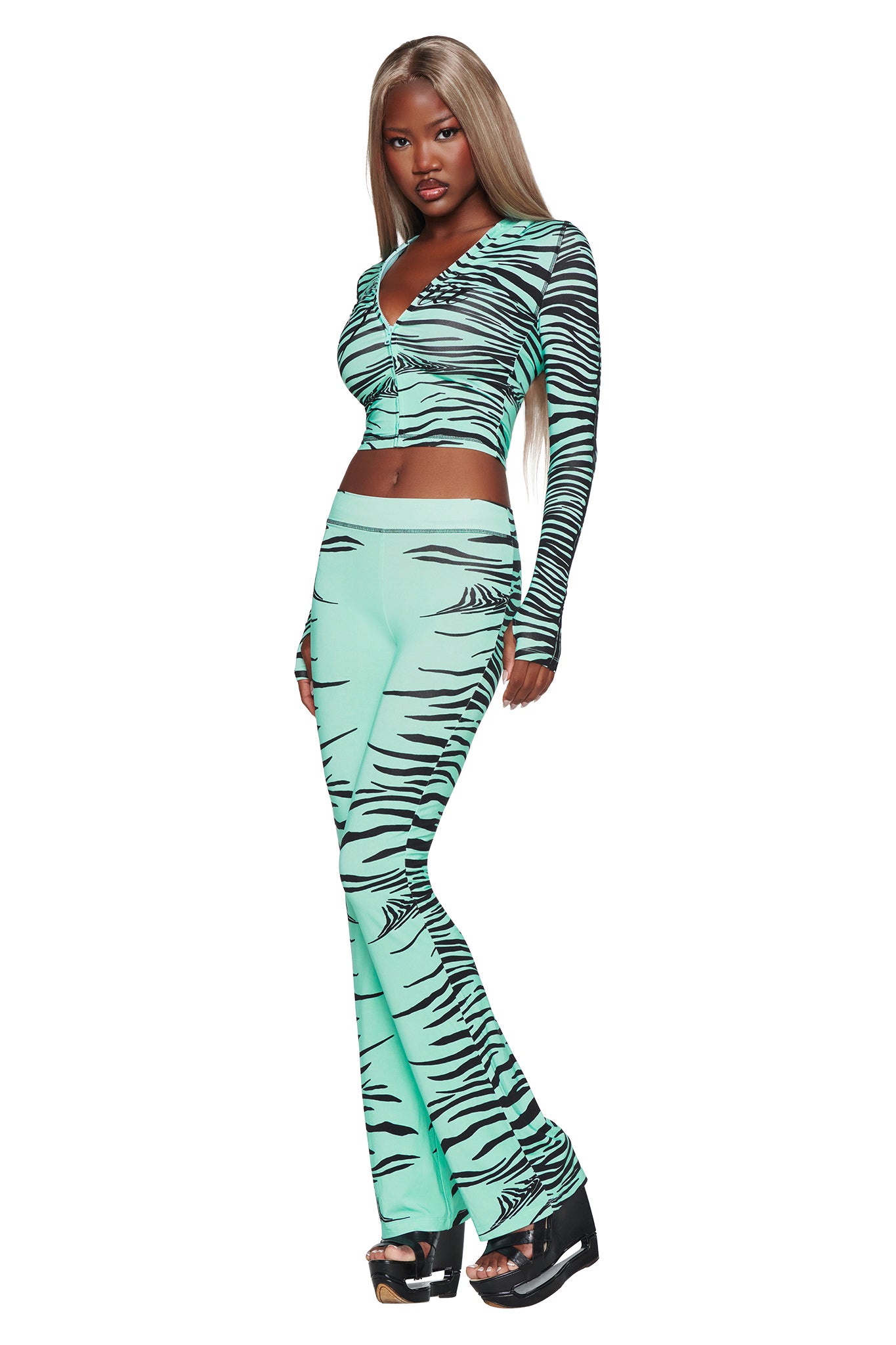 A woman models the I.AM.GIA BLARE TRACKPANT in Aqua Fine Line Zebra—a two-piece set featuring long sleeves and flared pants—styled with black platform sandals, against a white background.