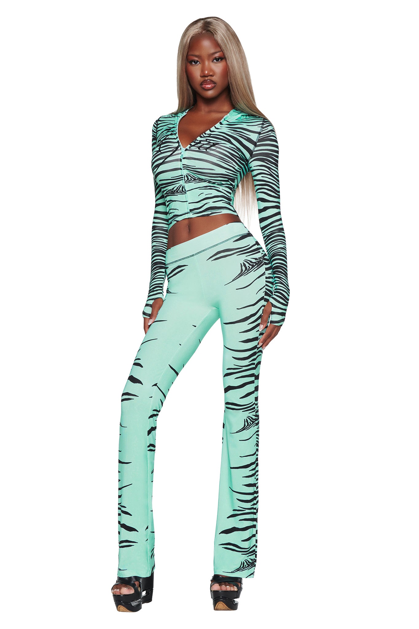 A woman with long blonde hair poses confidently against a white background, wearing the I.AM.GIA BLARE TRACKPANT in Aqua Fine Line Zebra—a light teal, zebra-print two-piece outfit featuring long sleeves and flared pants—paired with black platform heels.