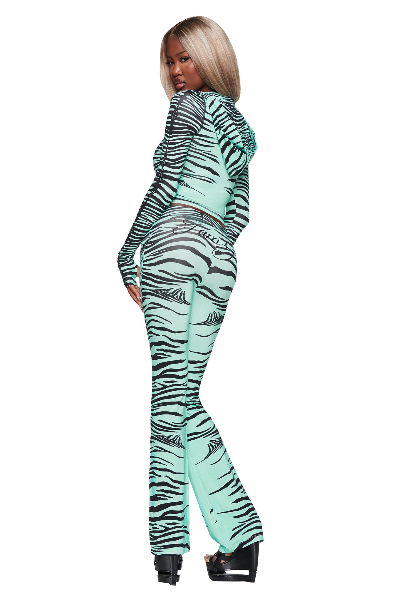 A woman with long blonde hair models the I.AM.GIA BLARE TRACKPANT in Aqua Fine Line Zebra, glancing over her shoulder as she stands tall on black platform heels against a crisp white background.