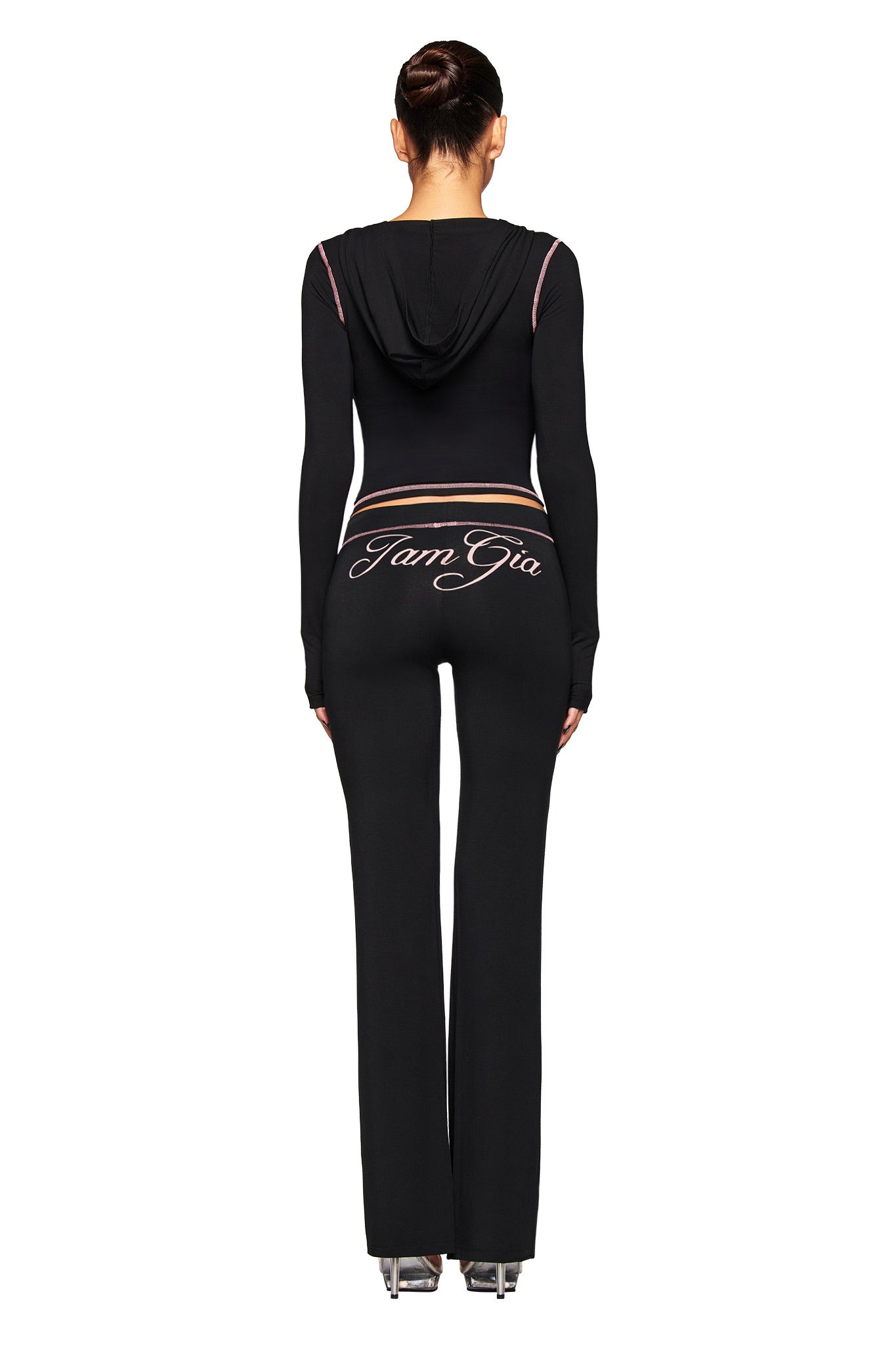 A woman, hair tied up, faces away wearing the I.AM.GIA BLARE HOODIE in black with matching black flared pants featuring "I.AM.GIA" cursive on the back, completed with chic high heels.