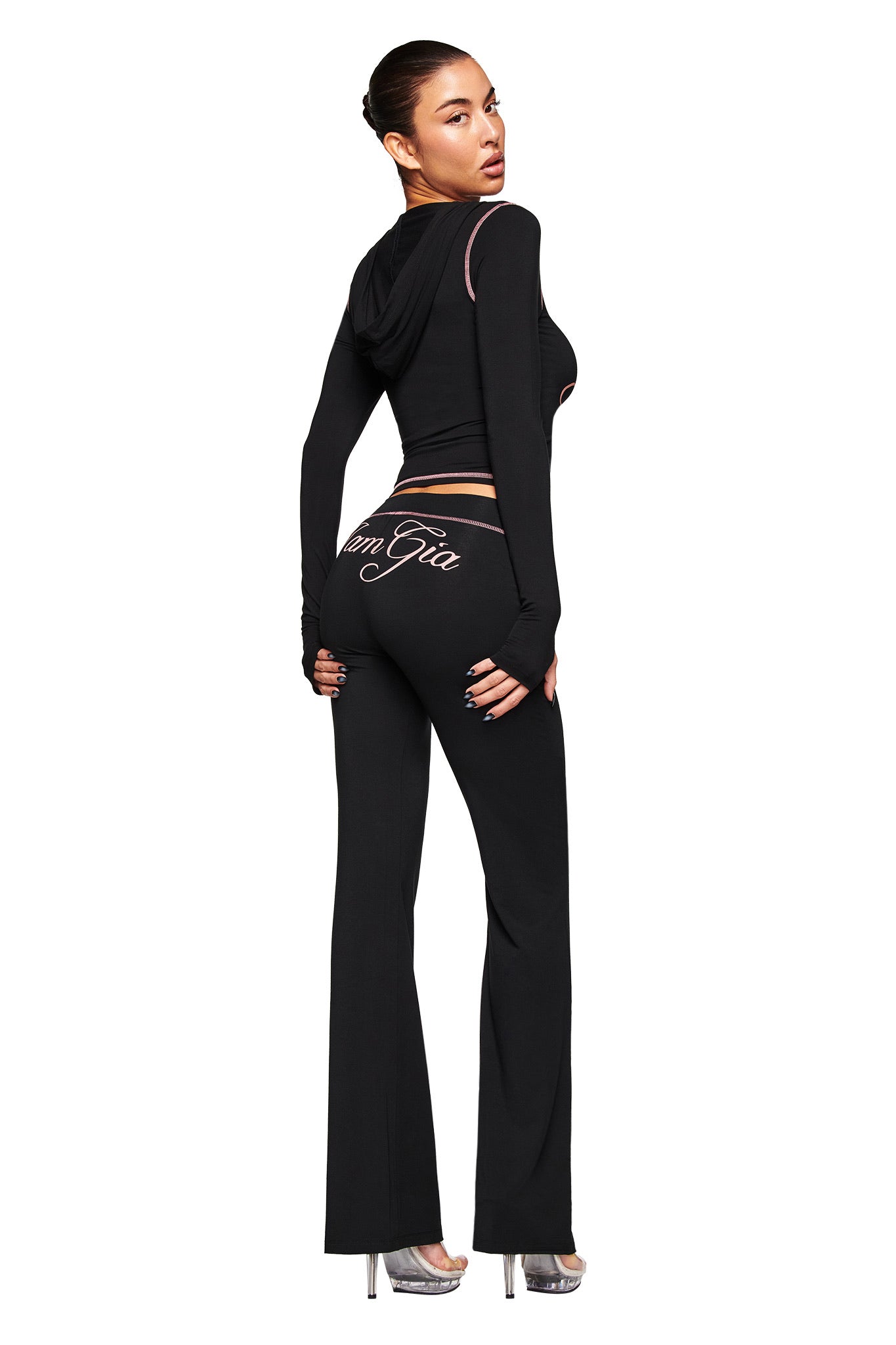 A woman poses in fitted black BLARE TRACKPANT by I.AM.GIA with "Von Gia" on the back, a matching hooded crop top, and silver high heels against a white background.