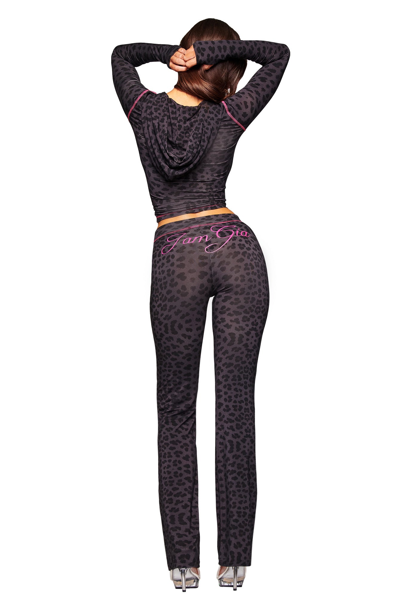 A woman stands with her back to the camera, arms raised, wearing the I.AM.GIA BLARE HOODIE - PANTHER and matching pants featuring “Fam Gia” in pink script, paired with silver high-heeled shoes.