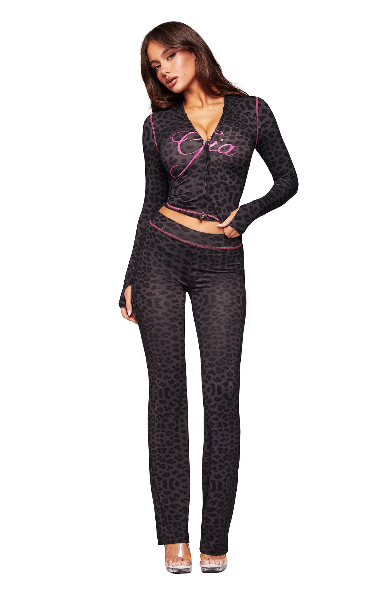 A woman models the I.AM.GIA BLARE HOODIE - PANTHER, a fitted black leopard print two-piece with long sleeves and flared pants. The top features "Gia" in pink script. She pairs it with clear heels against a white background.