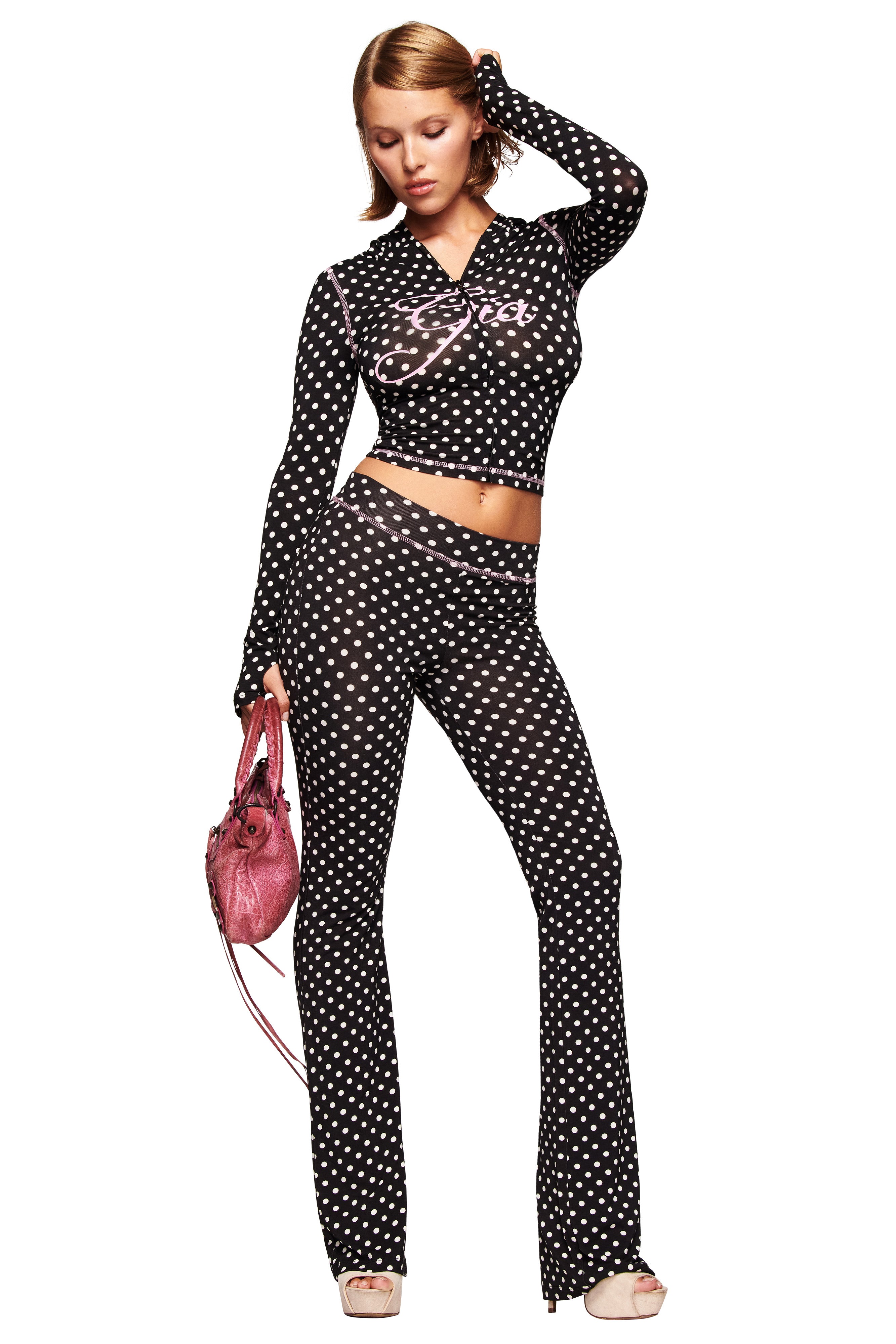 A woman models the I.AM.GIA BLARE TRACKPANT in black polka dot with a matching long-sleeve crop top, paired with high-heeled sandals and a pink handbag, all set against a white background.