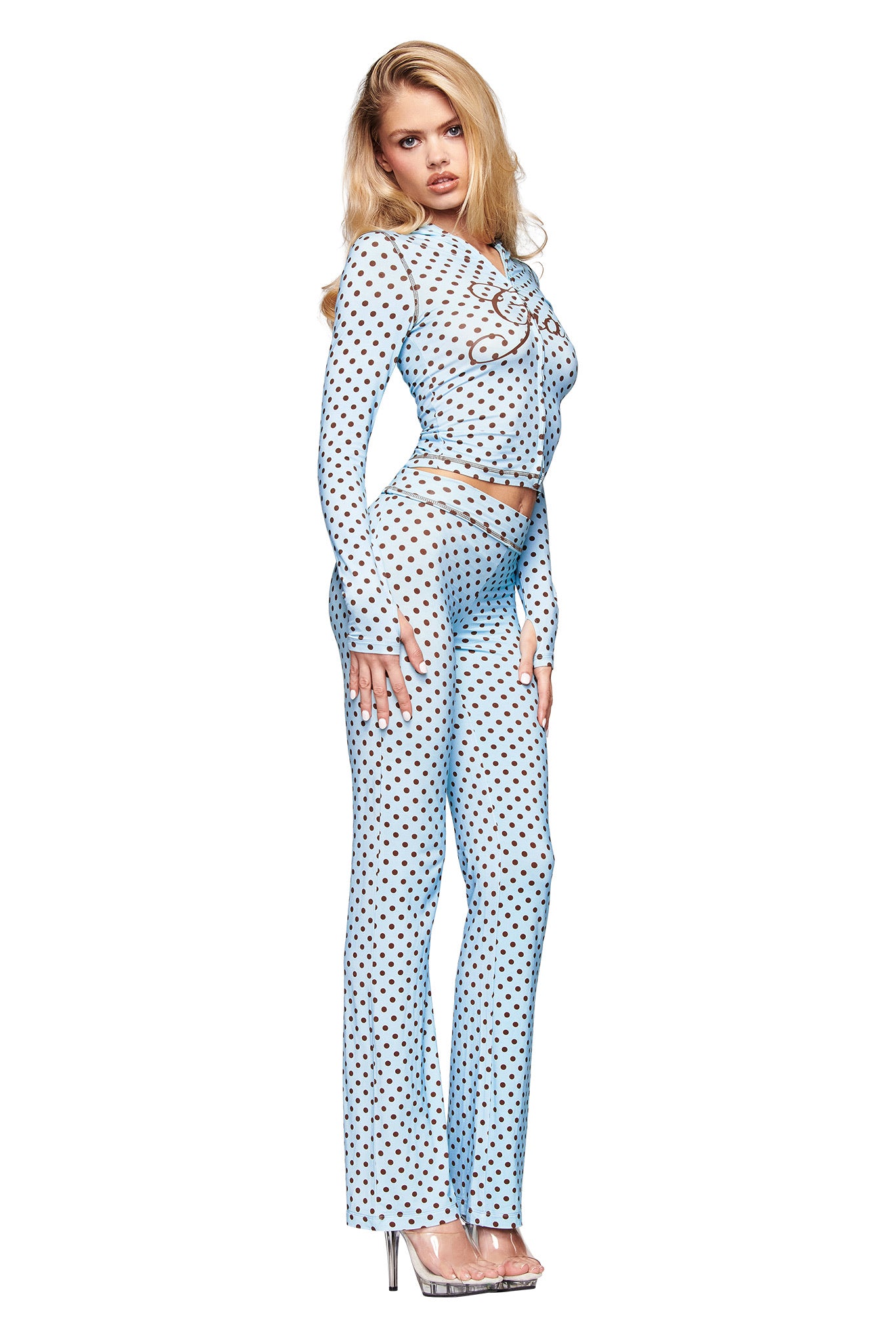 A woman with long blonde hair models the I.AM.GIA BLARE TRACKPANT in blue polka dot print, paired with a matching sheer long-sleeve top and clear high heels, set against a white background.