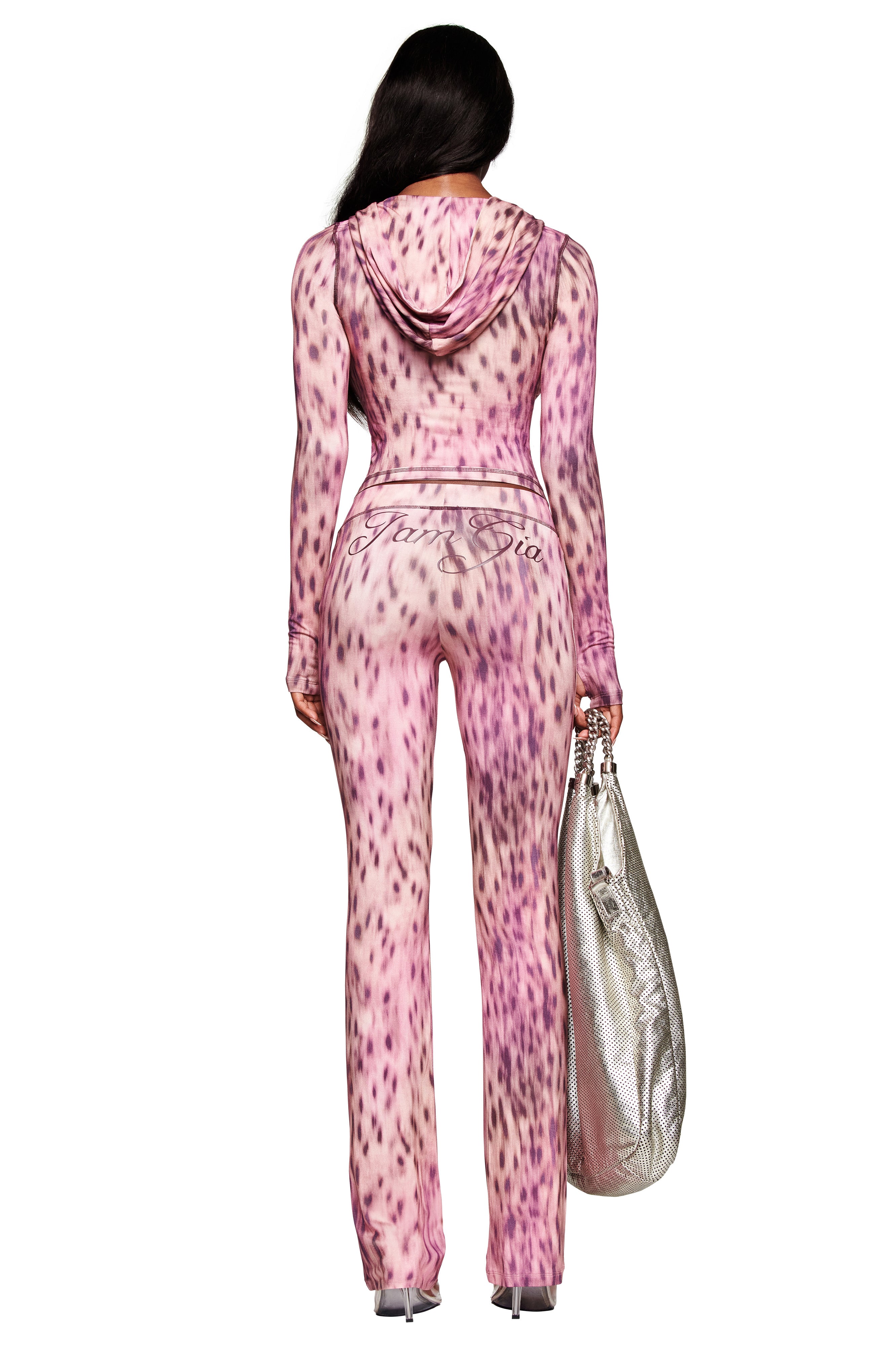 A woman faces away in the I.AM.GIA BLARE HOODIE - PINK BLURRED CHEETAH, a fitted bodycon style with a hood and cursive back detail. Her long dark hair falls over the hoodie as she holds a large silver bag and stands in heels.