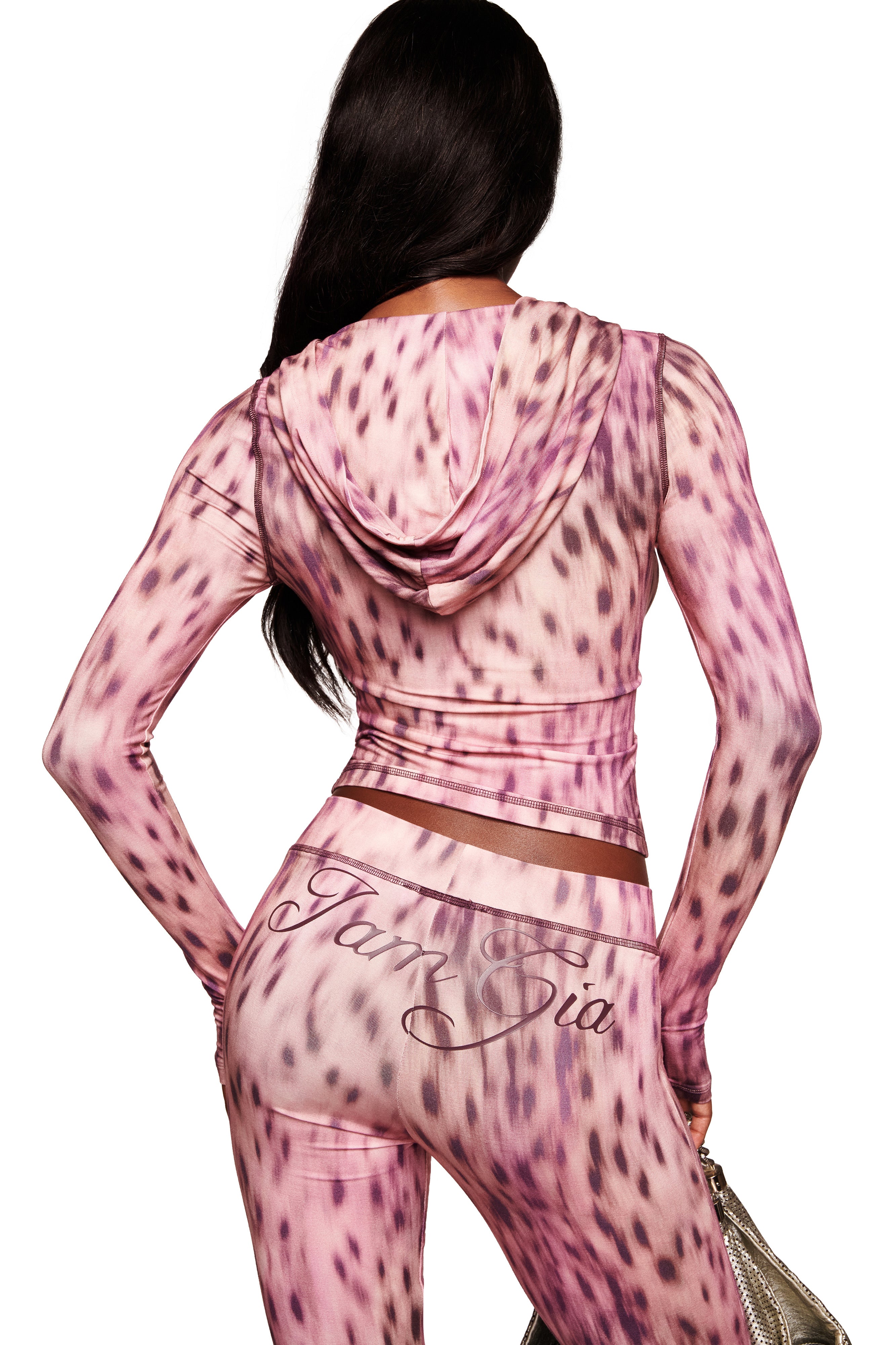 A woman with long dark hair, seen from behind, wears the I.AM.GIA BLARE HOODIE in Pink Blurred Cheetah print with matching leggings, both featuring the brand's logo. She carries a small shiny gold handbag.
