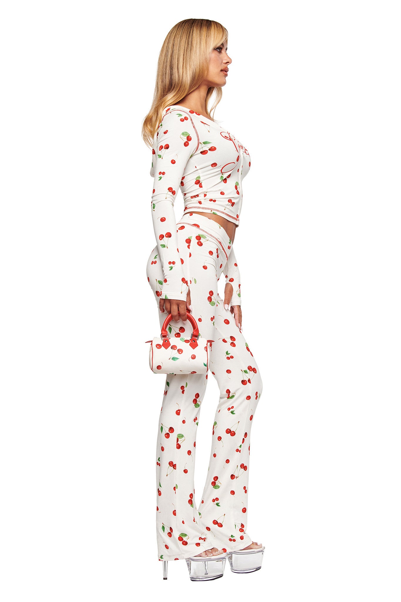 A woman stands sideways against a white background in I.AM.GIA's BLARE TRACKPANT - CHERRY ON TOP, paired with a white long-sleeve crop top, a matching small handbag, and clear platform heels.