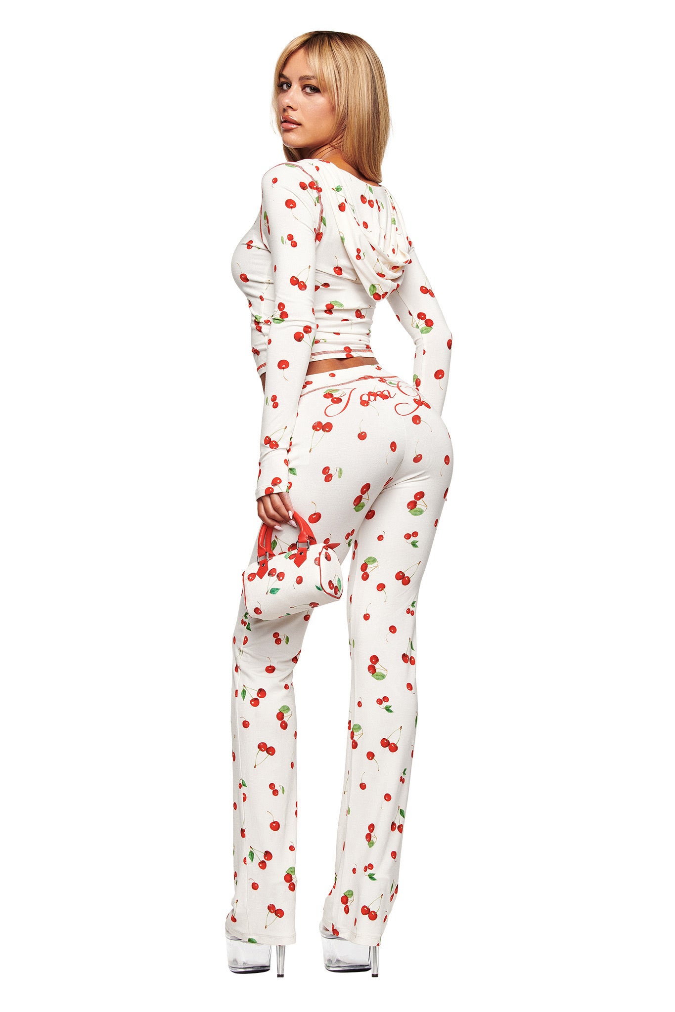 A person with long blonde hair in the fitted I.AM.GIA BLARE TRACKPANT - CHERRY ON TOP and clear heels stands facing away, looking over their shoulder, holding a small matching handbag by their side.