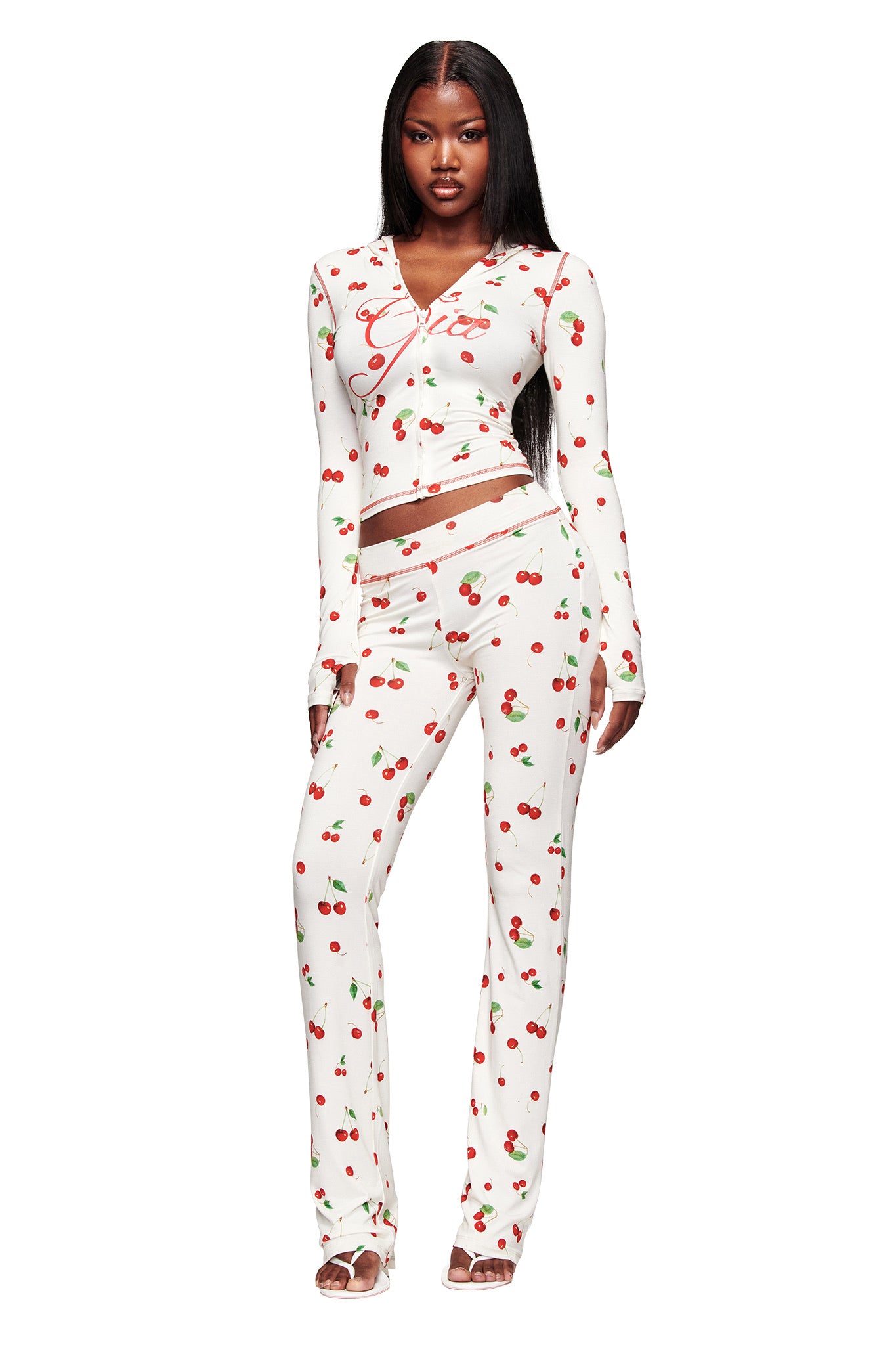 A woman with long straight hair wears I.AM.GIA's fitted BLARE TRACKPANT - CHERRY ON TOP and matching long-sleeve top, both featuring logo and cherry print. She stands forward in sandals with a neutral expression.