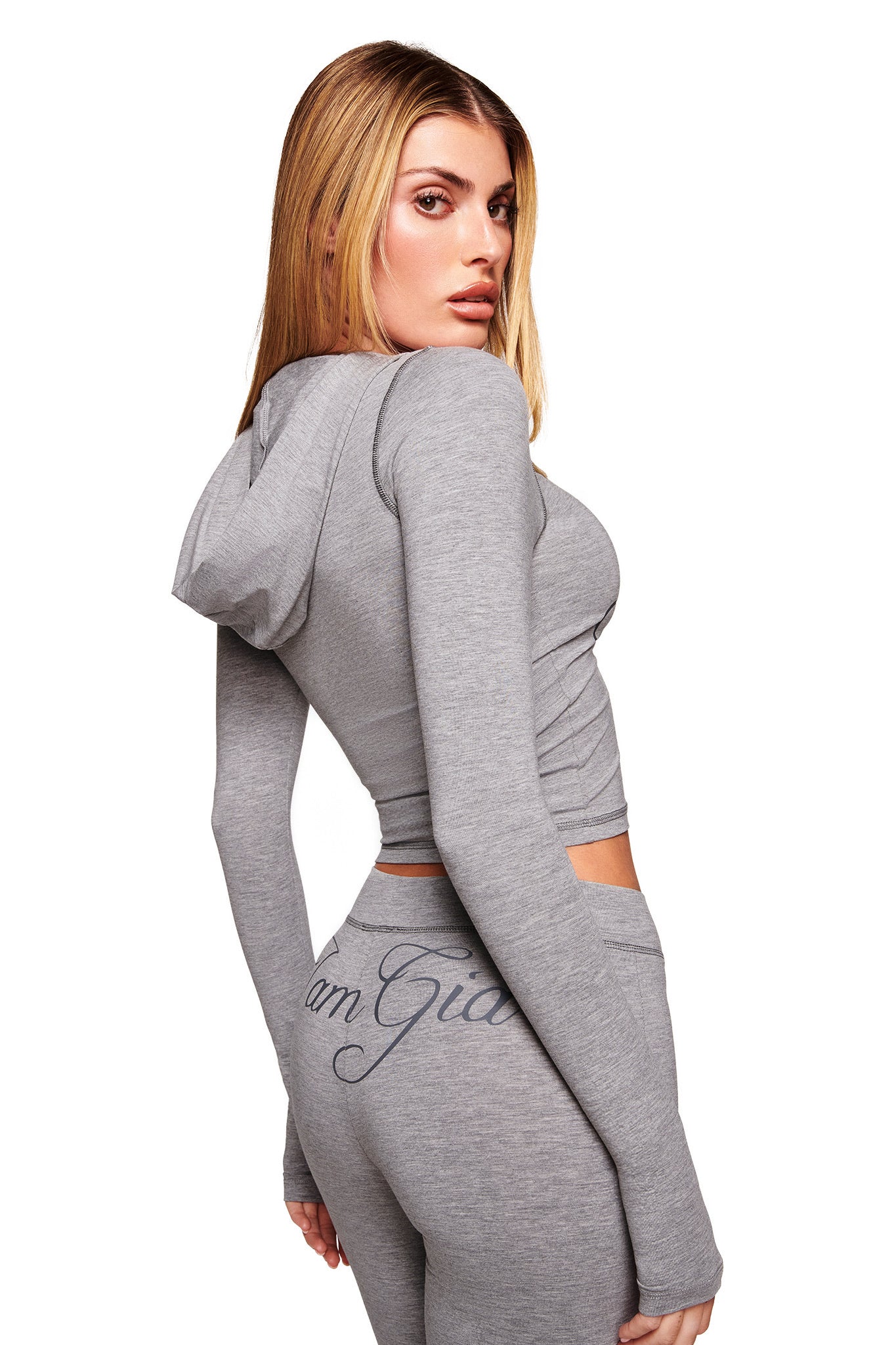 A woman with long blonde hair wears a light gray hooded crop top and I.AM.GIA BLARE JERSEY TRACKPANT in heather grey, "I am Gia" on the back, standing sideways and looking over her shoulder against a white background.