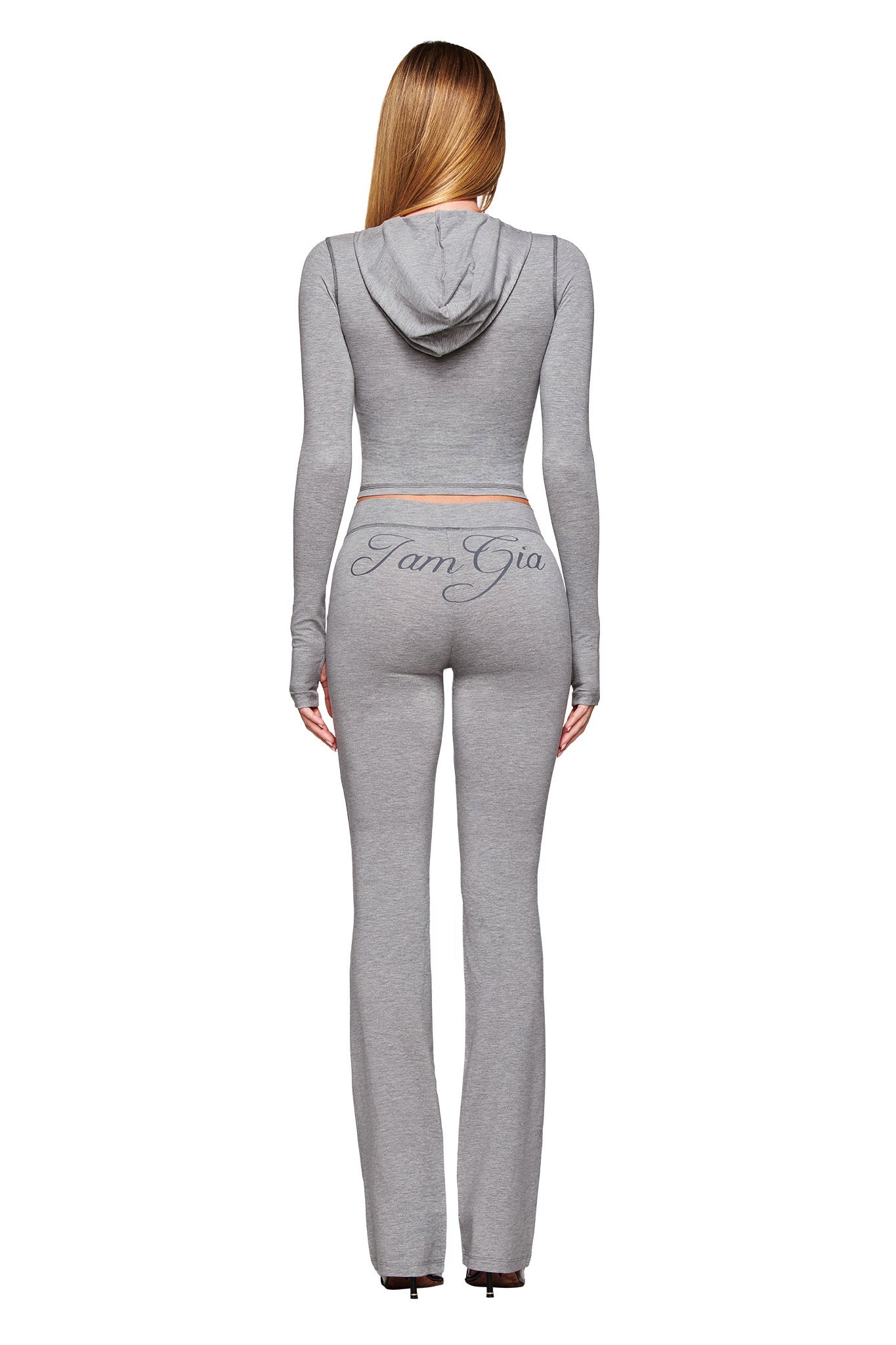 A woman with long straight hair, seen from behind, wears a fitted I.AM.GIA BLARE JERSEY HOODIE in heather grey with "I am Gia" across the back, paired with matching high-waisted pants.