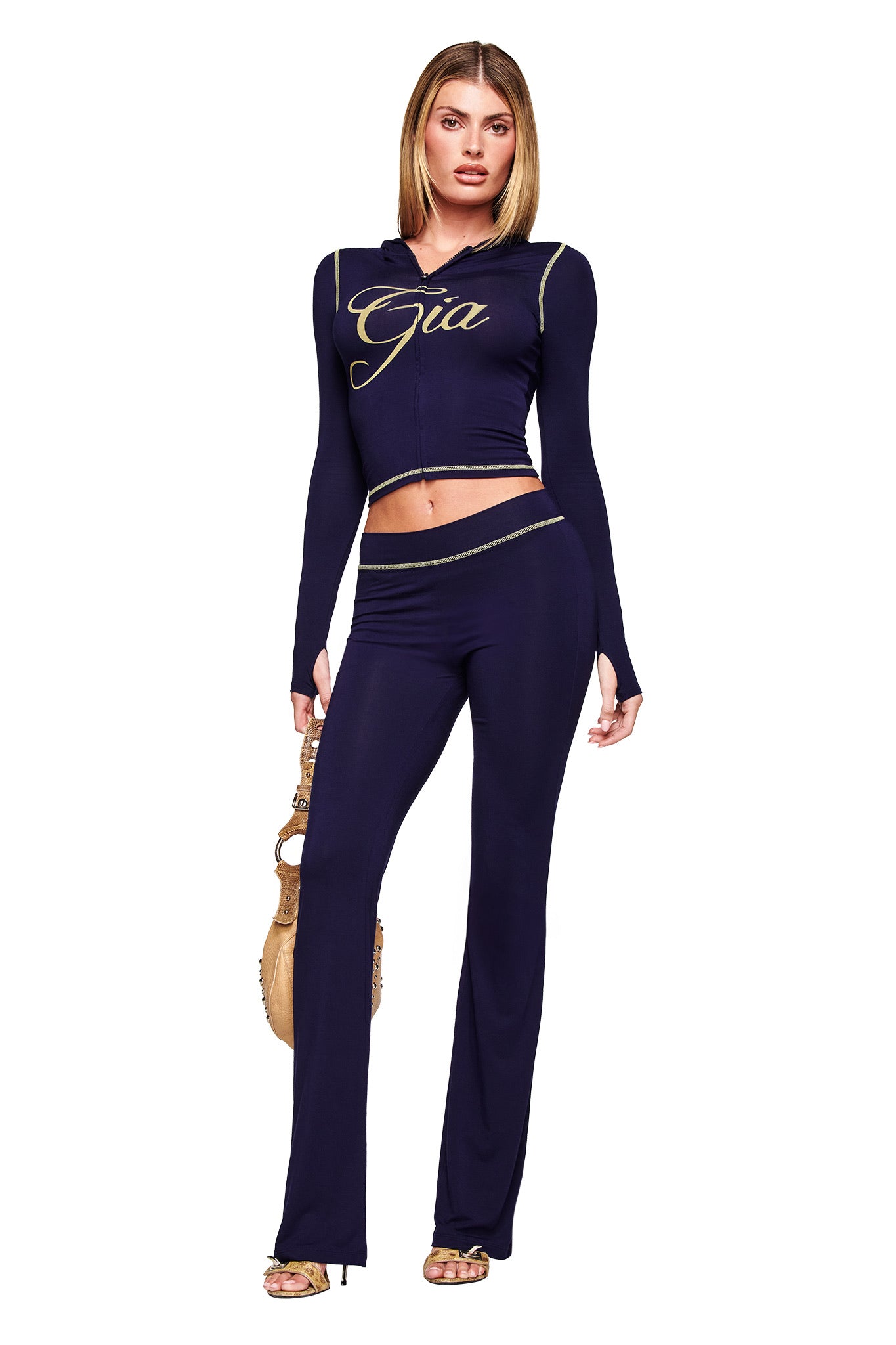 A woman models the I.AM.GIA BLARE HOODIE - NAVY set, featuring a hooded crop top with gold trim and matching flared pants. She holds a tan handbag, pairs the look with gold heels, and poses against a white background.