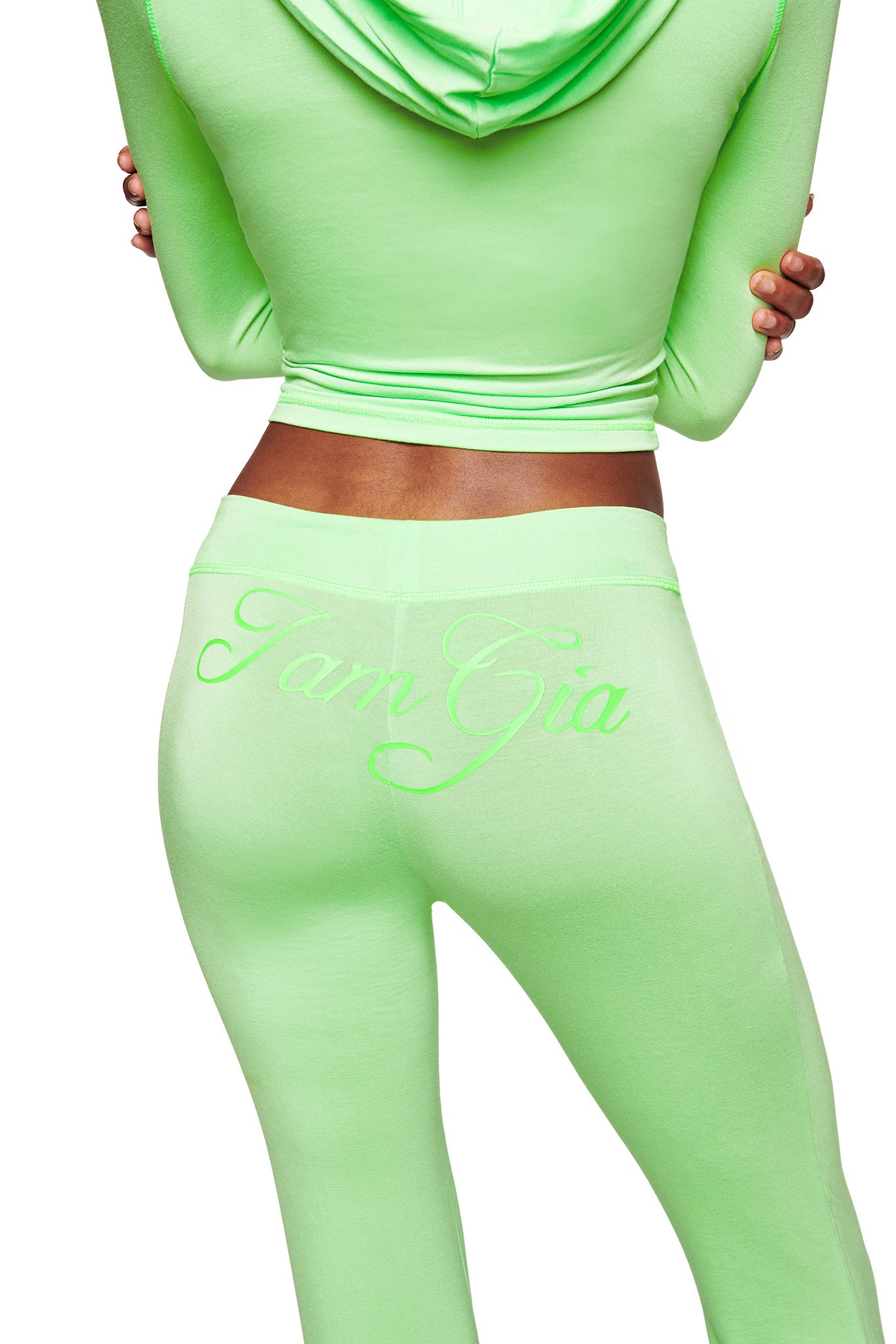 A person is shown from behind wearing the BLARE TRACKPANT - NEON GREEN by I.AM.GIA, a fitted, low-rise set with flared legs and matching neon green "I.AM.GIA" embroidery on the back. Their hands are positioned behind their back.