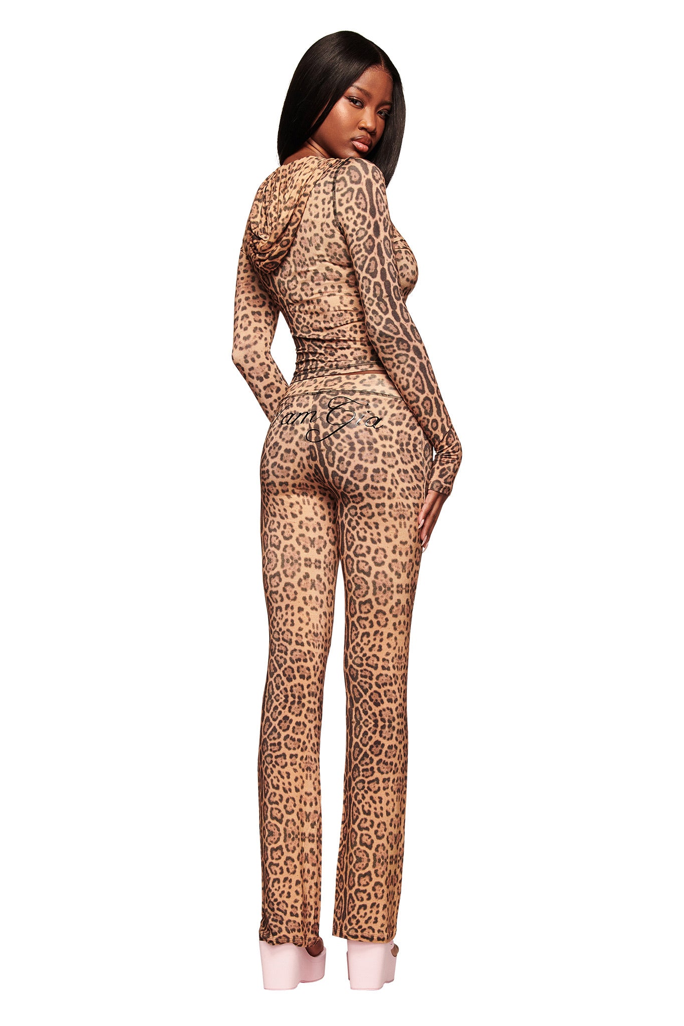 A woman poses in I.AM.GIA’s BLARE TRACKPANT - PANTHERA, a fitted, long-sleeved hooded jumpsuit with a brown and black cheetah print. She looks over her shoulder in light pink heels against a white background.