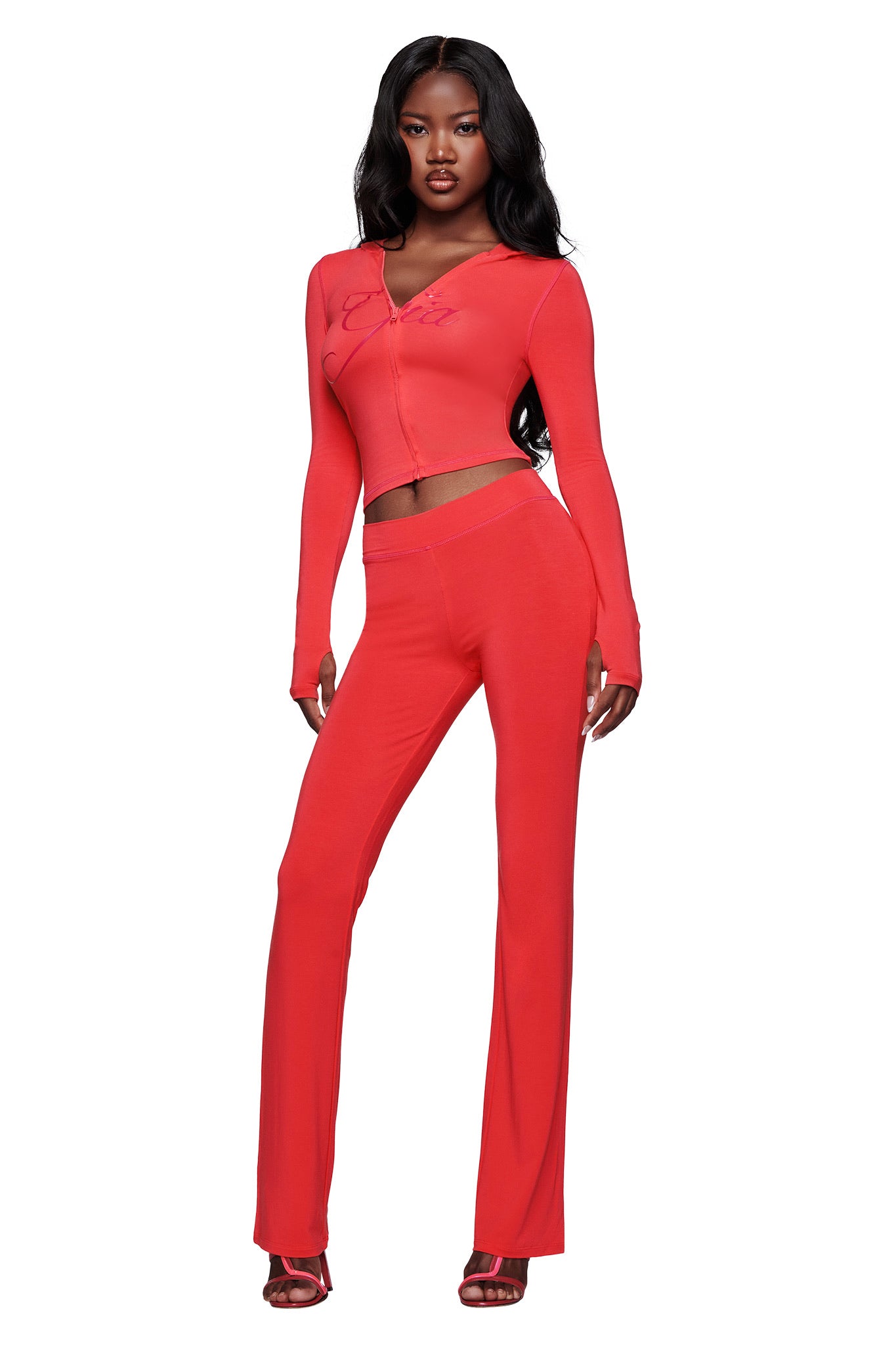 A woman stands confidently in a fitted, long-sleeve red top and matching I.AM.GIA BLARE TRACKPANT - PAPAYA flared pants, finished with red heels against a white background.