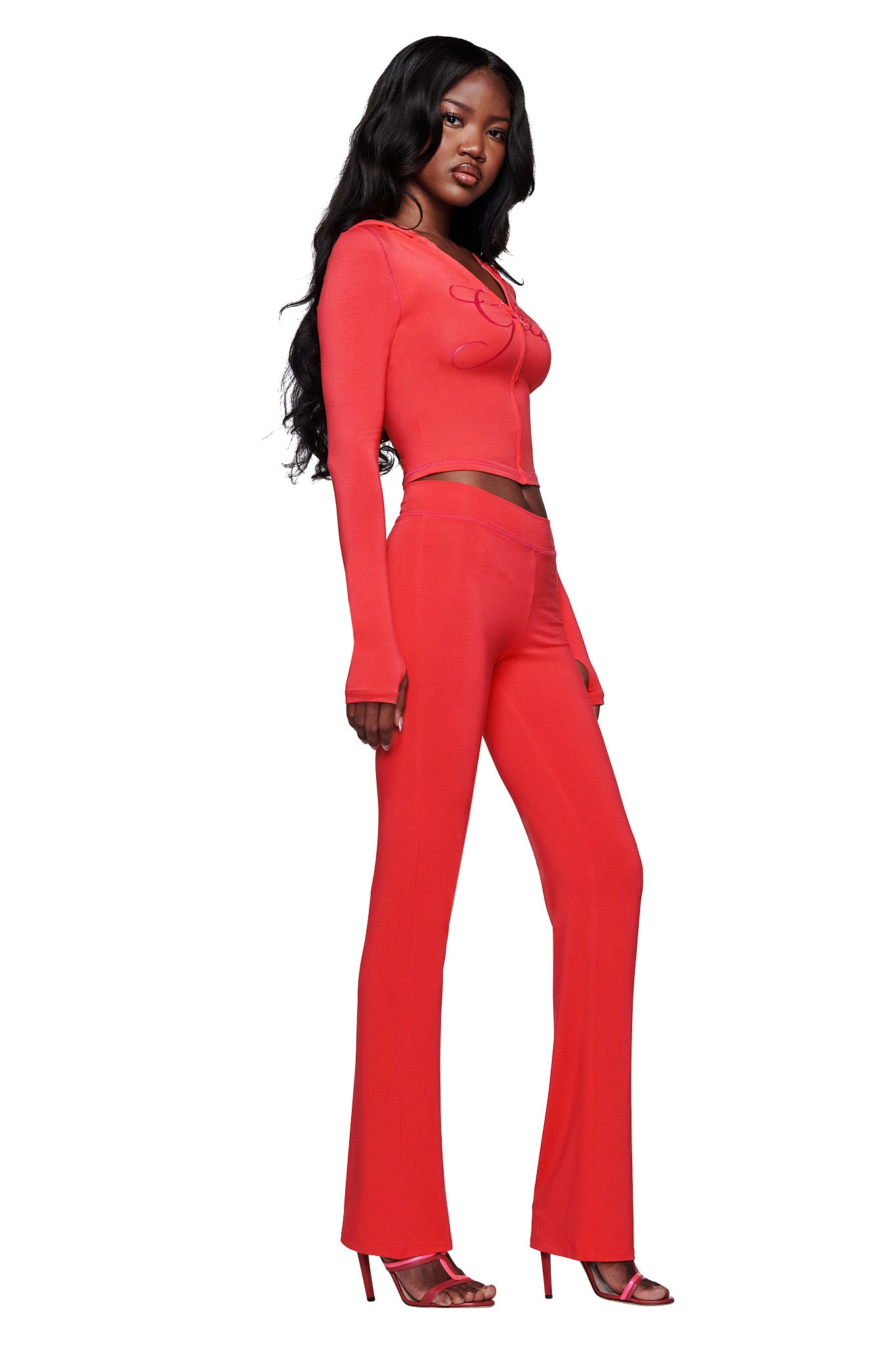 A woman with long black hair stands sideways in a fitted bright red crop top, I.AM.GIA BLARE TRACKPANT - PAPAYA flared leg pants, and red high-heeled sandals against a white background.