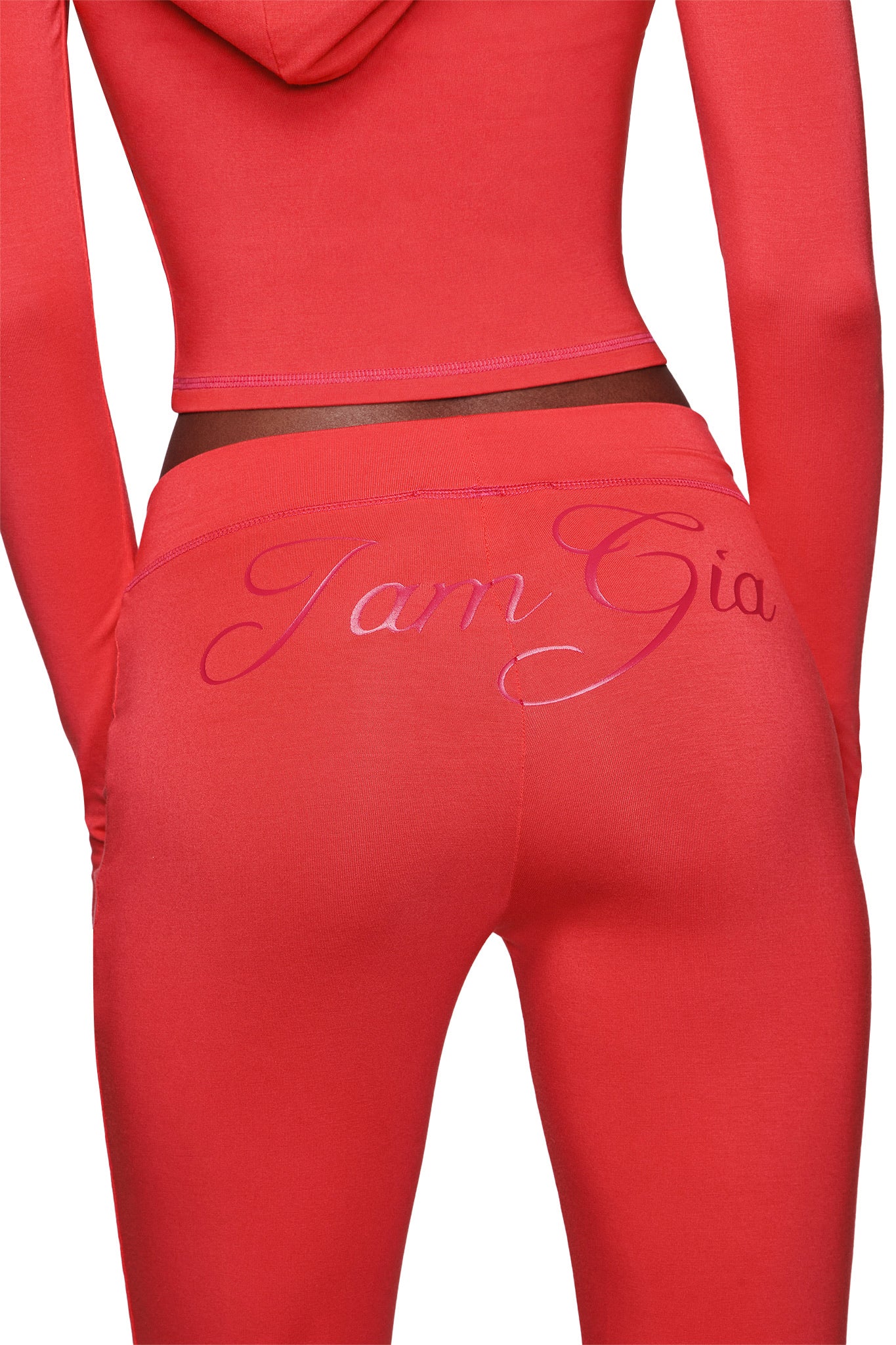 A person wears the I.AM.GIA BLARE TRACKPANT - PAPAYA, featuring "I.AM.GIA" in shiny script on the back. The fitted, mid-rise trackpants are cropped from mid-back to mid-thigh and have a trendy flared leg style.