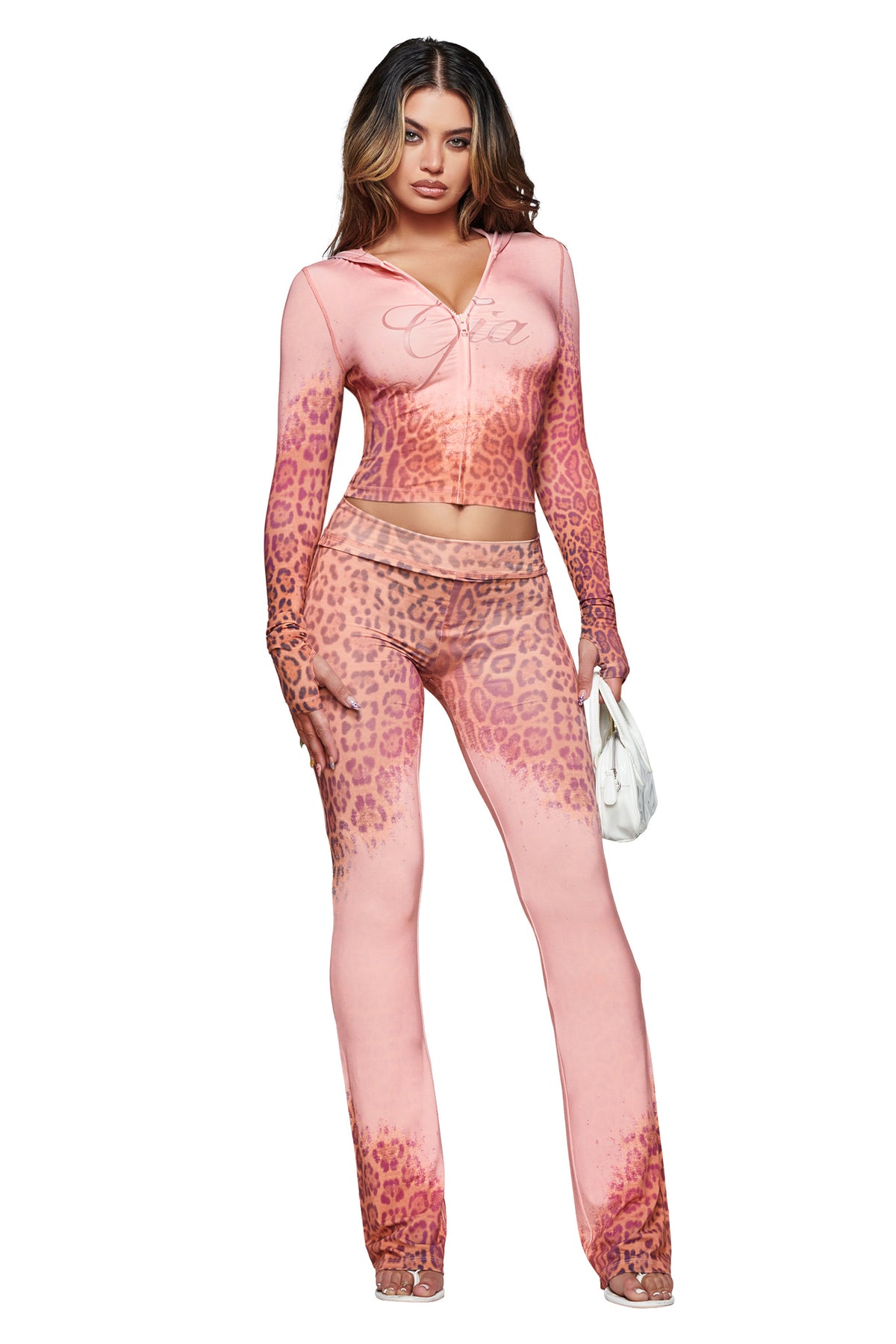 A woman poses against a white background wearing the I.AM.GIA BLARE TRACKPANT in Pink Faded Panthera, styled as part of a matching leopard-print two-piece outfit with long sleeves and flared pants, and carries a small white handbag.