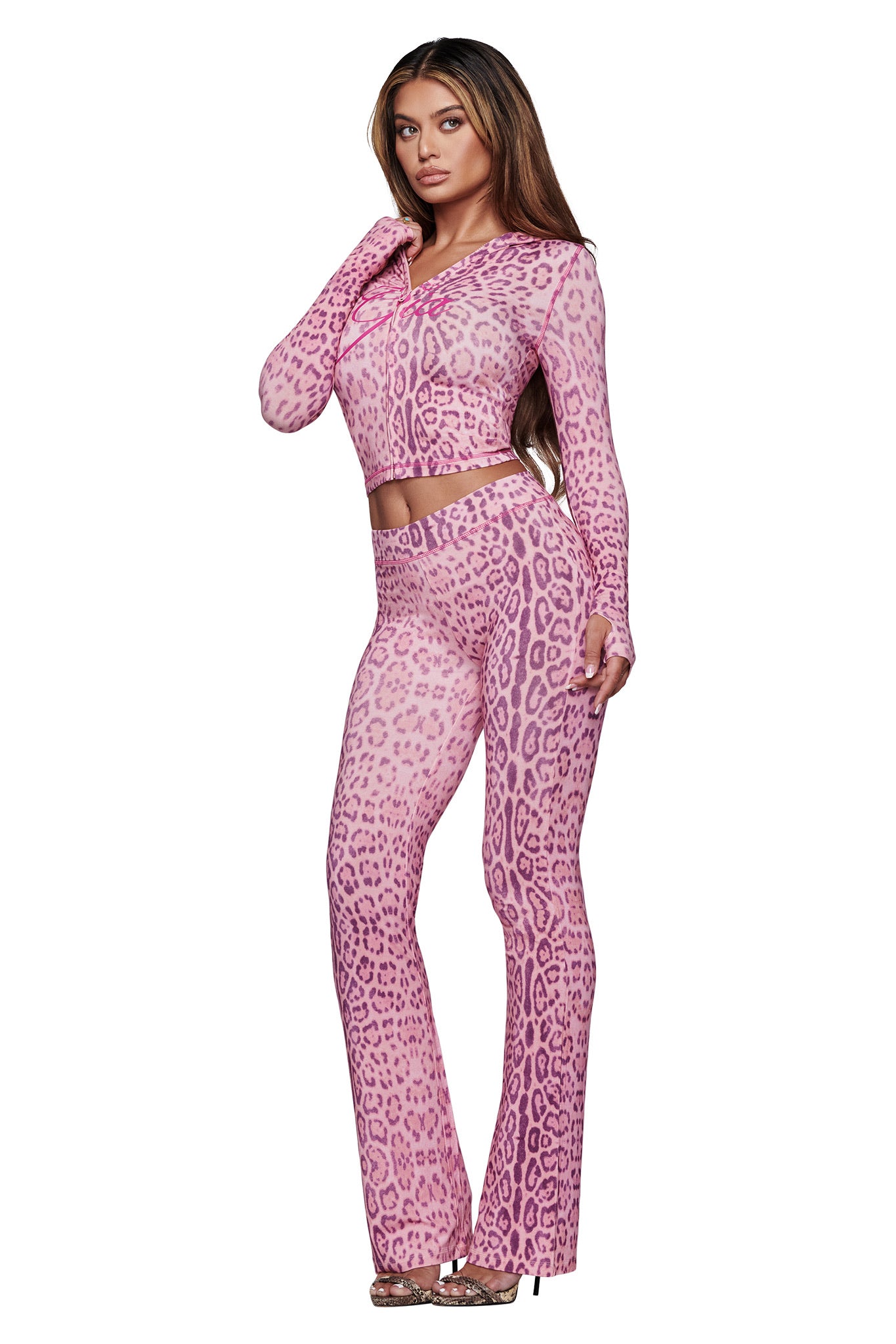 A woman poses in the I.AM.GIA BLARE TRACKPANT - PINK PANTHERA, featuring a leopard-print, flared-leg silhouette, paired with the matching crop top and high heels against a white background.