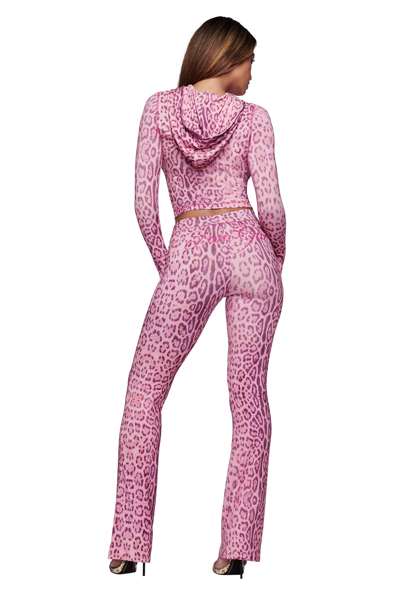 A woman faces away, wearing I.AM.GIA's BLARE TRACKPANT - PINK PANTHERA, a fitted flared-leg trackpant with matching hooded top in a bold leopard print, styled with high heels.