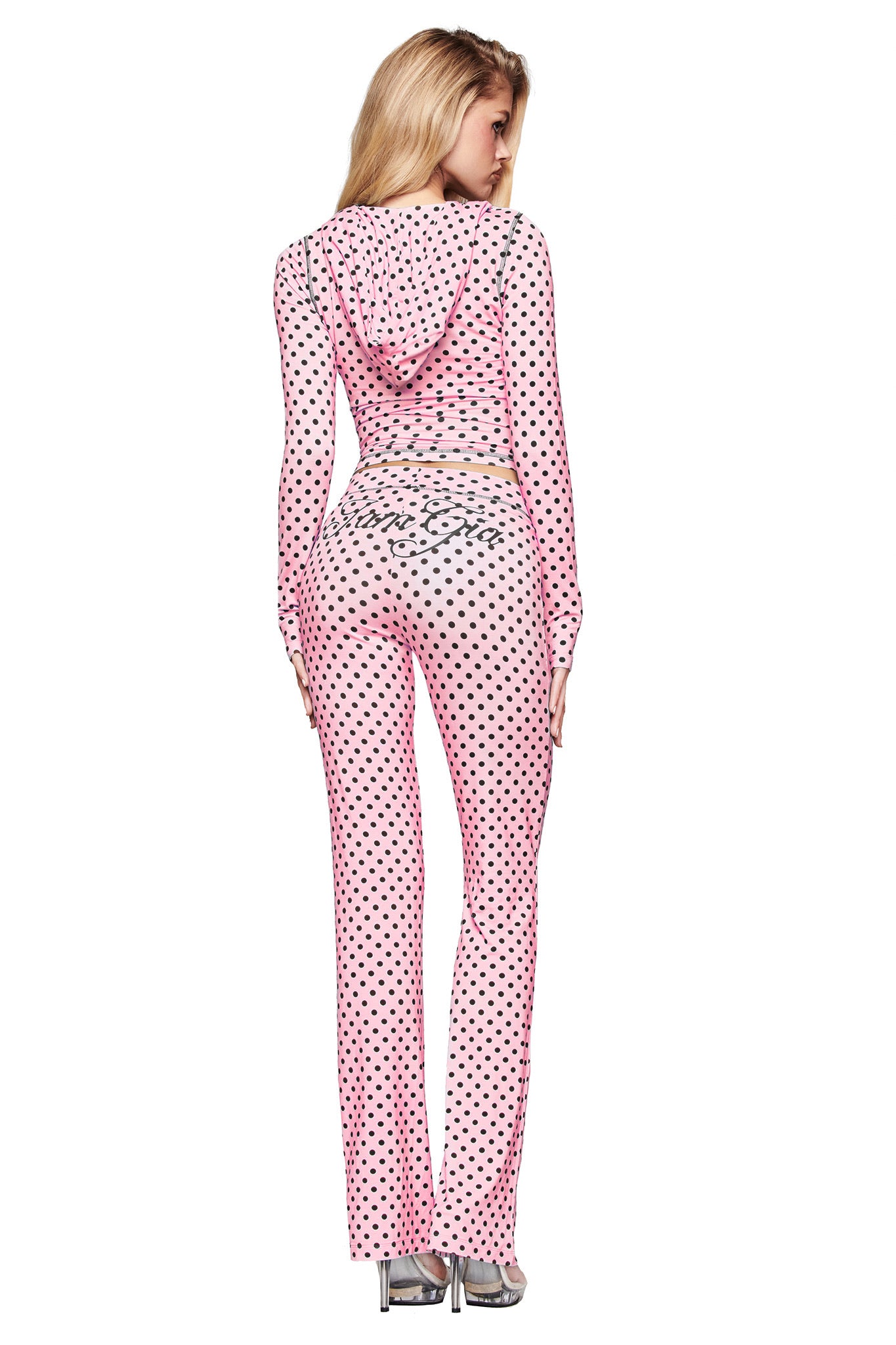 A woman with long blonde hair showcases the I.AM.GIA BLARE HOODIE - PINK POLKA DOT set, featuring a chic hooded mesh outfit with flared pants. She faces away to highlight the stylish back design.