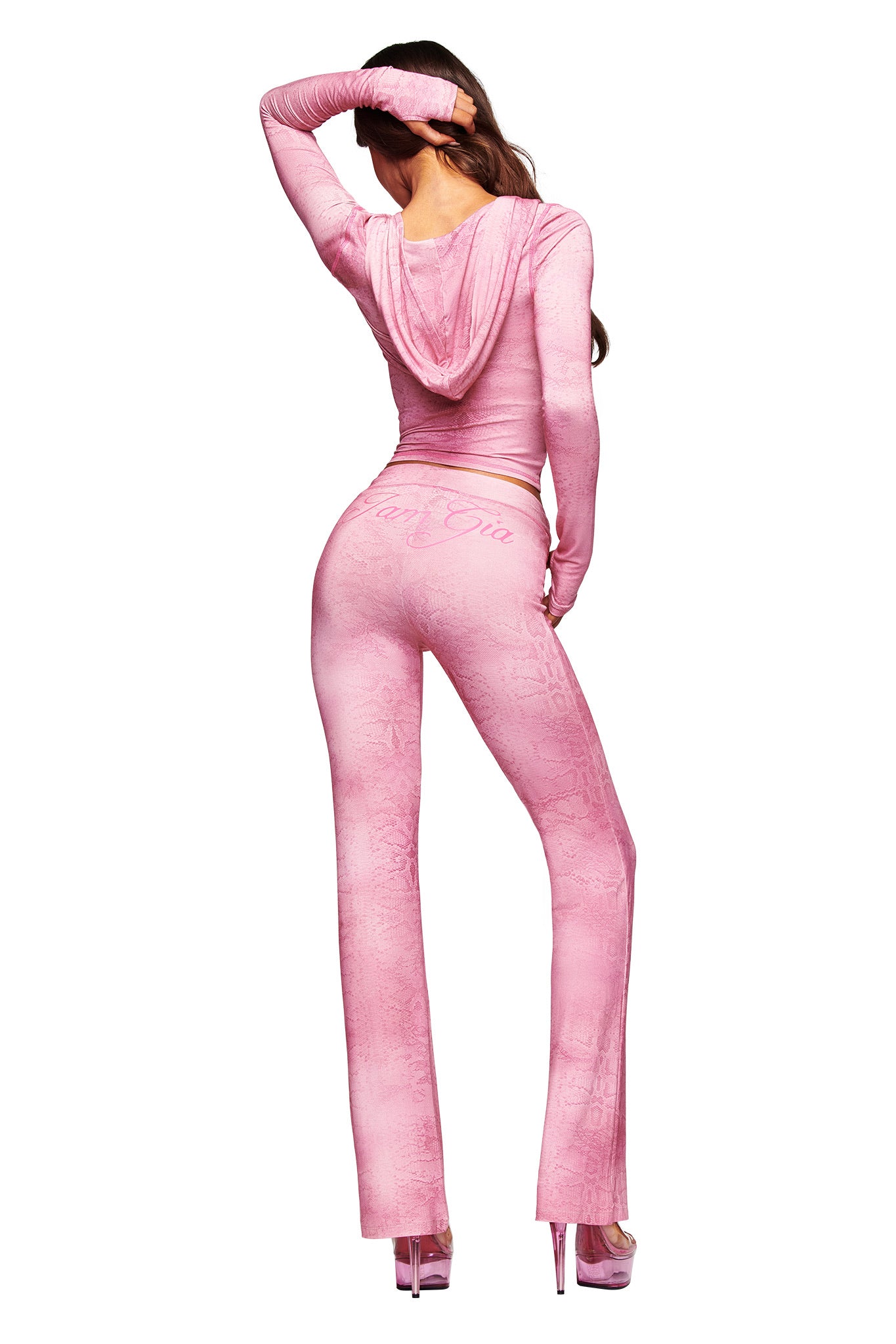 A woman with long brown hair poses facing away, wearing I.AM.GIA's BLARE TRACKPANT - LIGHT PINK SERPENTINE set with a hood and flared pants, paired with high pink platform heels, against a white background.