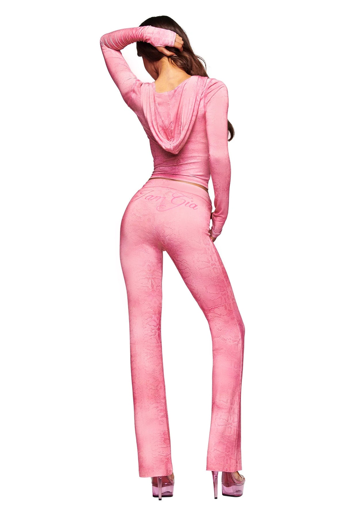 A woman stands facing away, wearing the I.AM.GIA BLARE TRACKPANT in Light Pink Serpentine—with a fitted silhouette, flared leg, and long sleeves—one arm bent as her hand rests on her head.