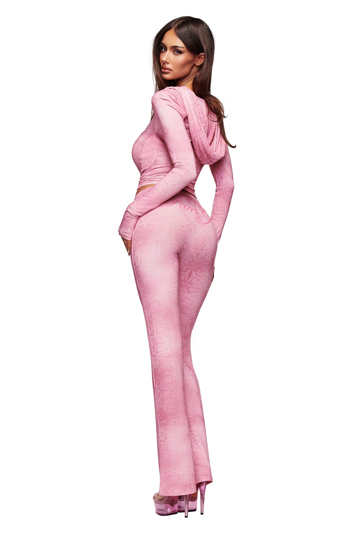 A woman in a fitted I.AM.GIA hooded top and matching BLARE TRACKPANT - LIGHT PINK SERPENTINE stands in high-heeled sandals, looking over her shoulder against a plain white background.