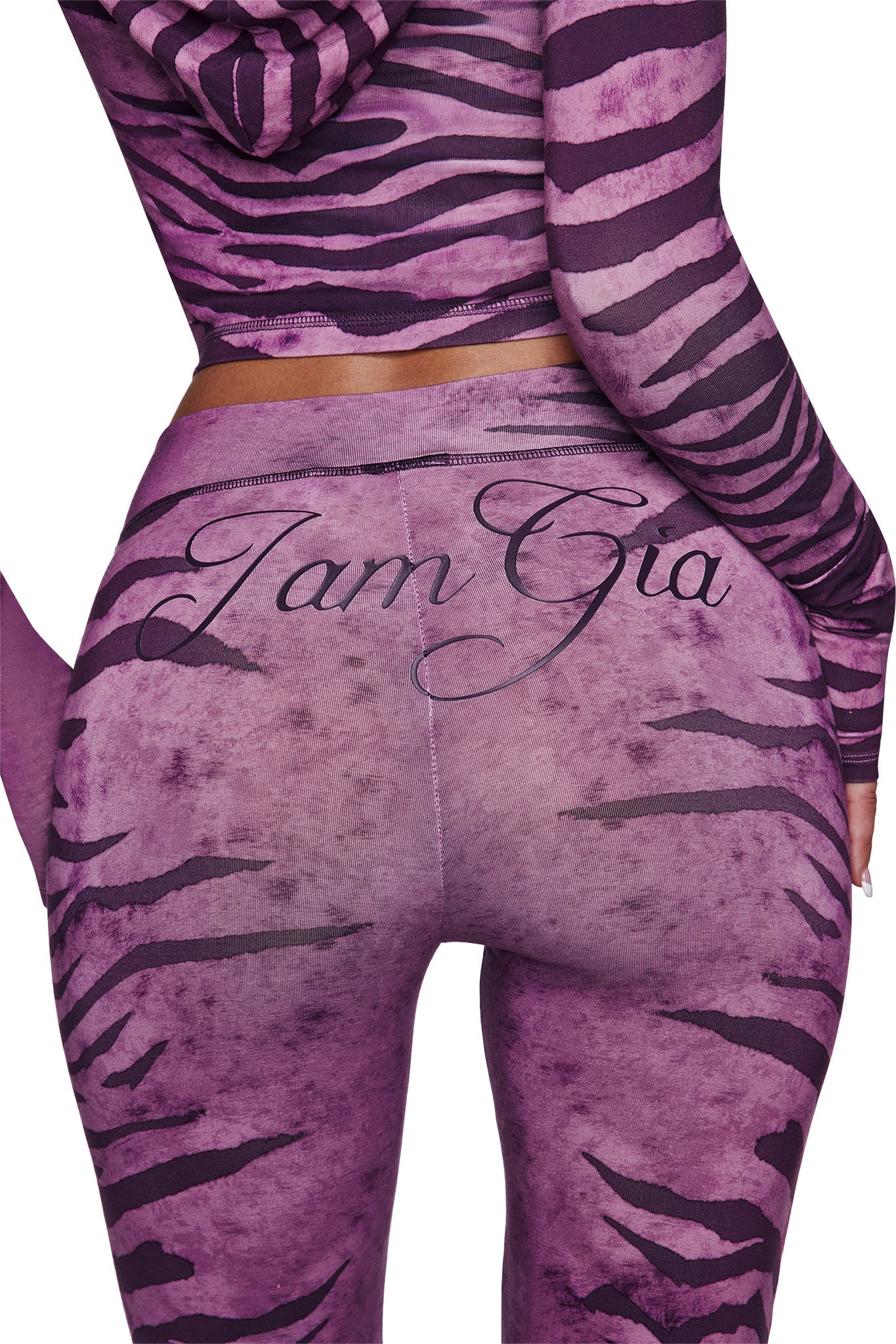 A person wearing I.AM.GIA's BLARE TRACKPANT - PLUM WILD SIDE, with flared legs, purple and black tiger stripes, and "I am Gia" in cursive across the back. Photo is cropped to show only the lower back and upper legs.