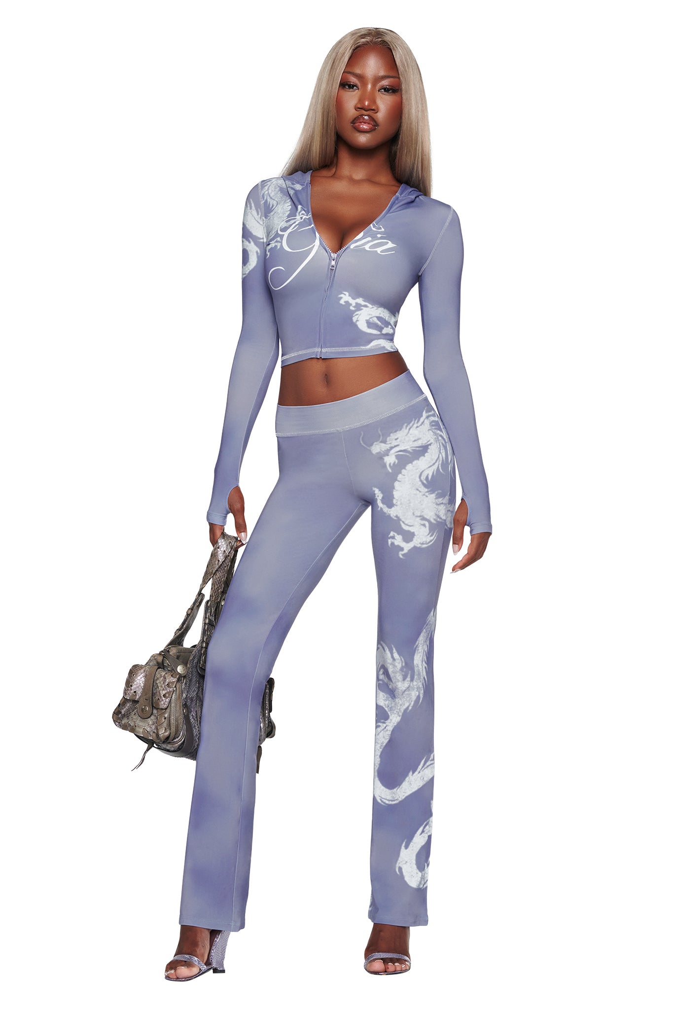 A woman poses confidently in the I.AM.GIA BLARE TRACKPANT - STEEL BLUE DRAGON, a two-piece outfit featuring bold white swirling patterns, paired with a silver handbag and heeled sandals.