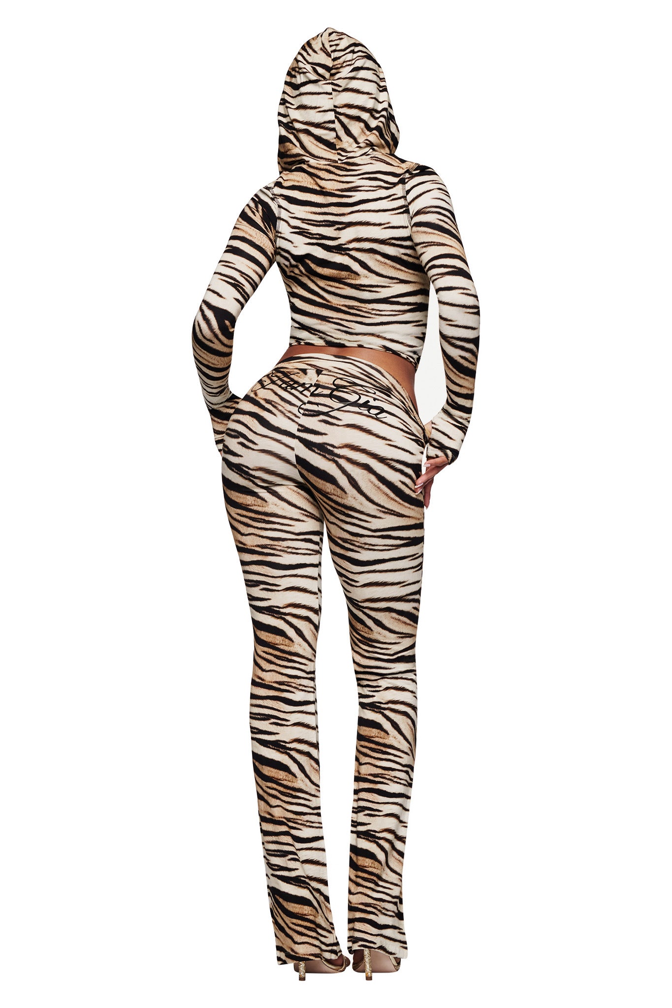 A person faces away wearing the I.AM.GIA BLARE TRACKPANT - LIGHT TIGRESS, featuring beige, black, and white tiger stripes in a fitted bamboo trackpant style with a hooded top and flared legs, paired with high heels.