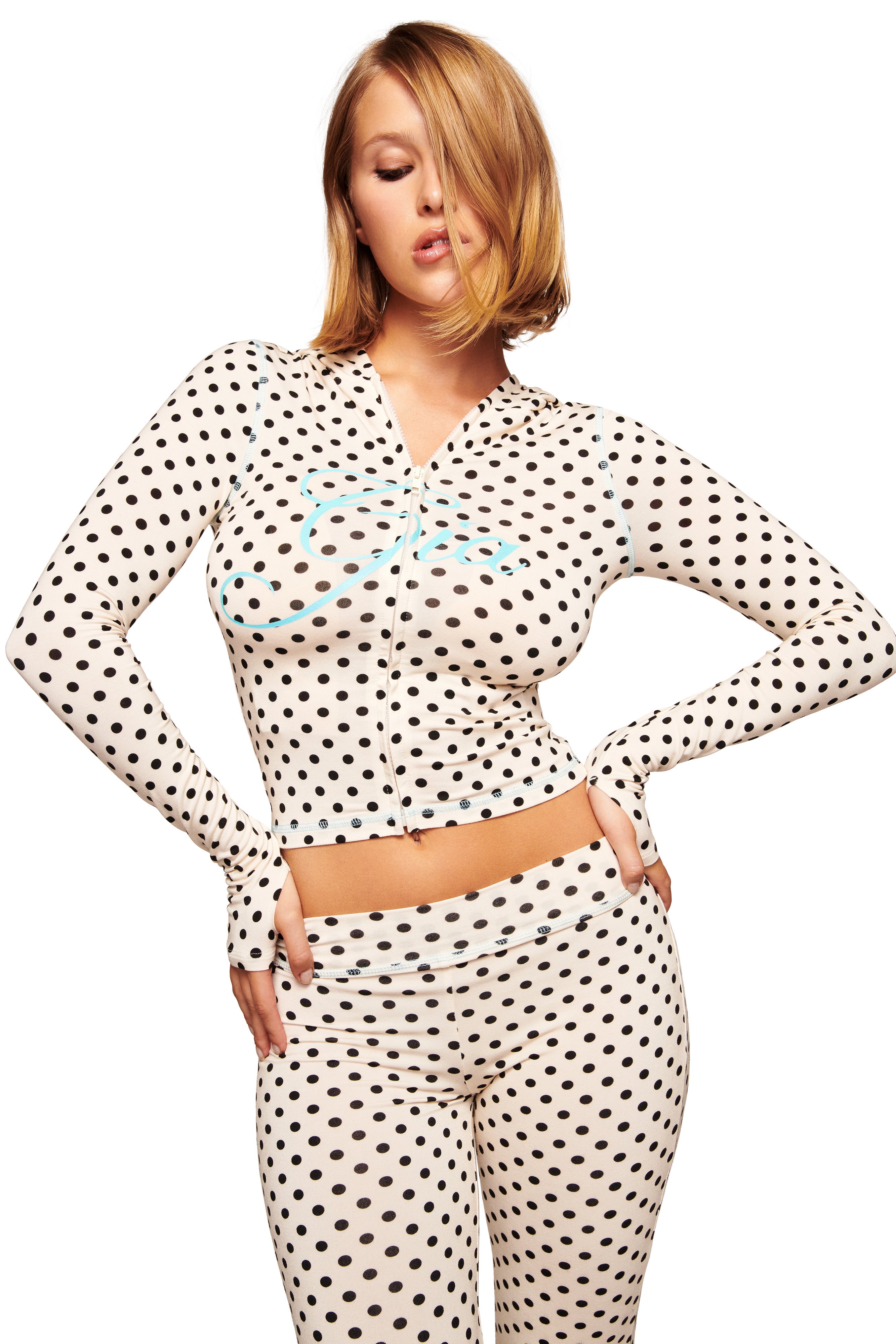 A woman poses in the I.AM.GIA BLARE HOODIE - CREAM POLKA DOT, a fitted, long-sleeve hoodie with white polka dots. She stands with her left hand on her hip, hair partially over one eye, against a white background.