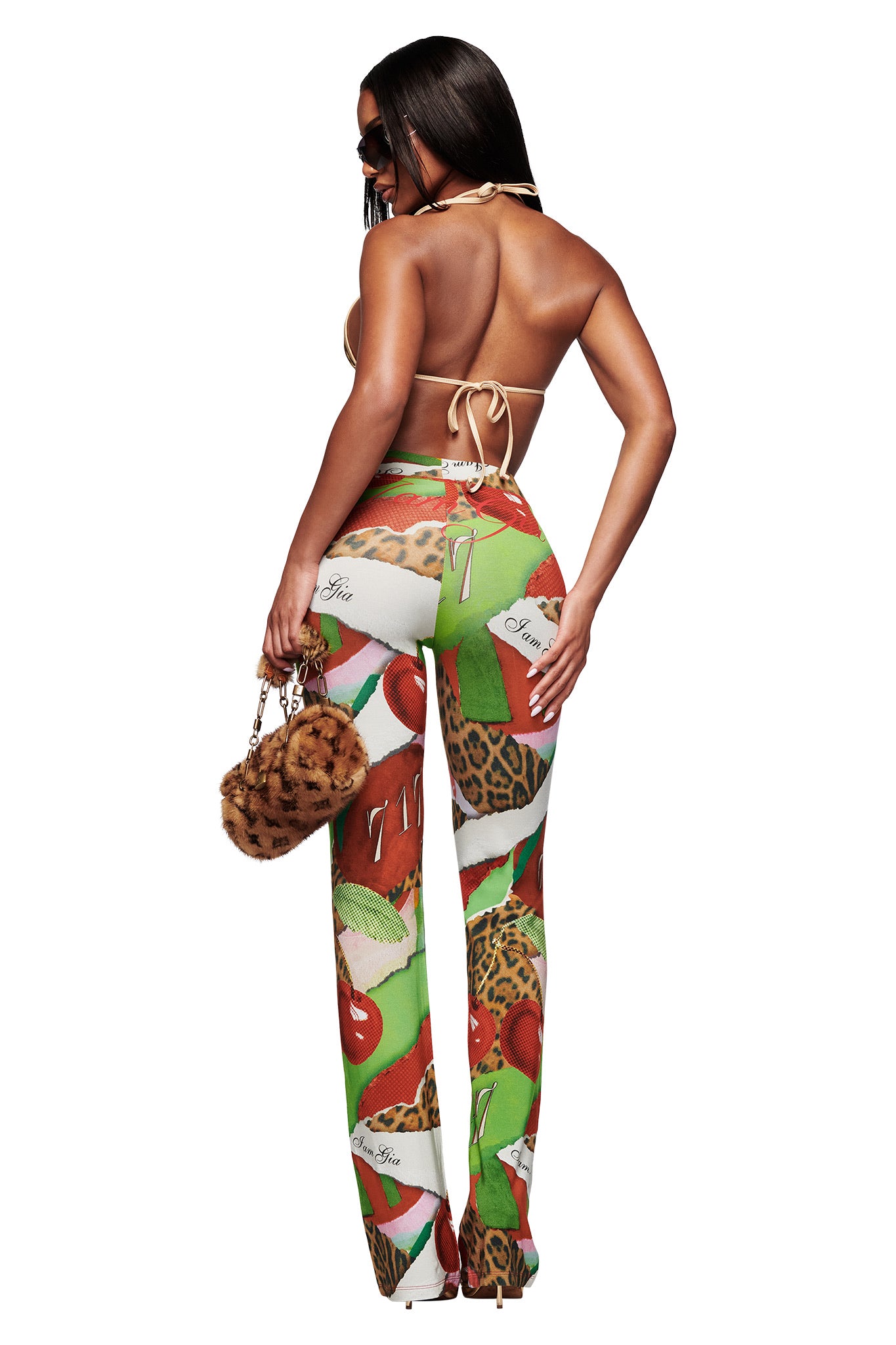 A woman faces away, wearing the I.AM.GIA BLARE TRACKPANT in Wild Cherry with a backless halter top, sunglasses, long straight hair down her back, and carrying a small fur bag.