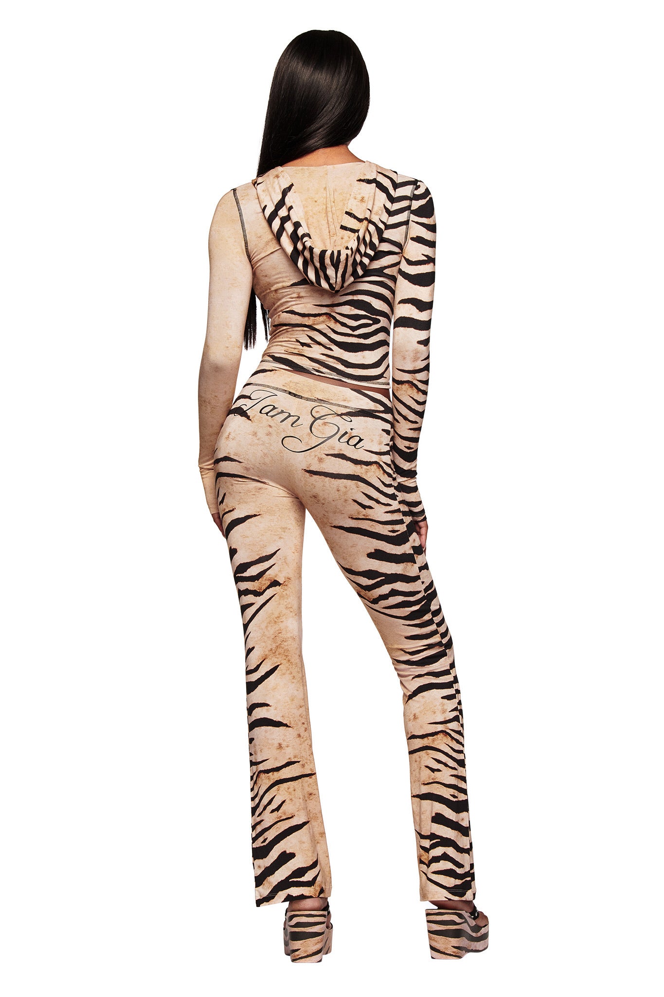 A woman with long dark hair faces away, wearing the I.AM.GIA BLARE HOODIE in Wild Side print and platform shoes. "I.AM.GIA" is printed on the back of the beige and black bamboo-fabric hoodie.