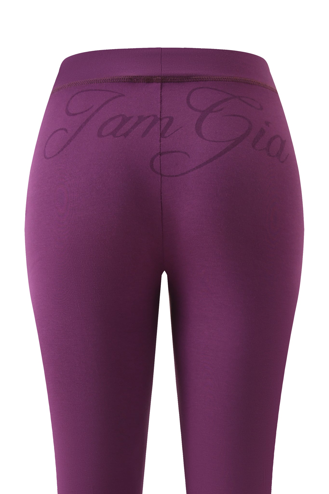 The BLARE TRACKPANT - PLUM by I.AM.GIA is shown from the back, with “I.AM.GIA” in cursive across the low-rise waistband. The smooth, form-fitting leggings appear in a purple plum shade.