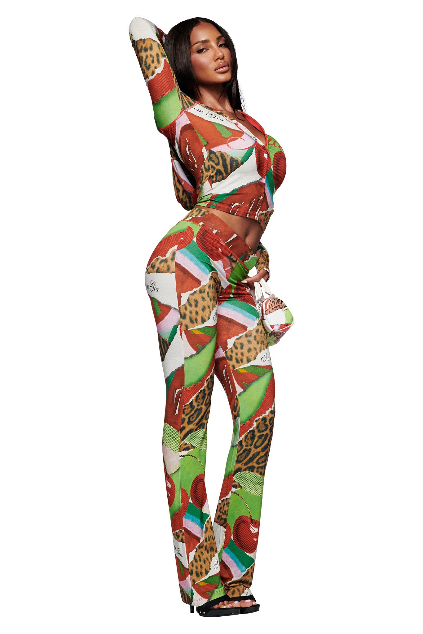 A woman poses confidently in the I.AM.GIA BLARE HOODIE - WILD CHERRY, a vibrant, form-fitting piece featuring bold prints of wild cherry, watermelon, leopard spots, and geometric shapes on white. She stands in high heels against a plain backdrop.