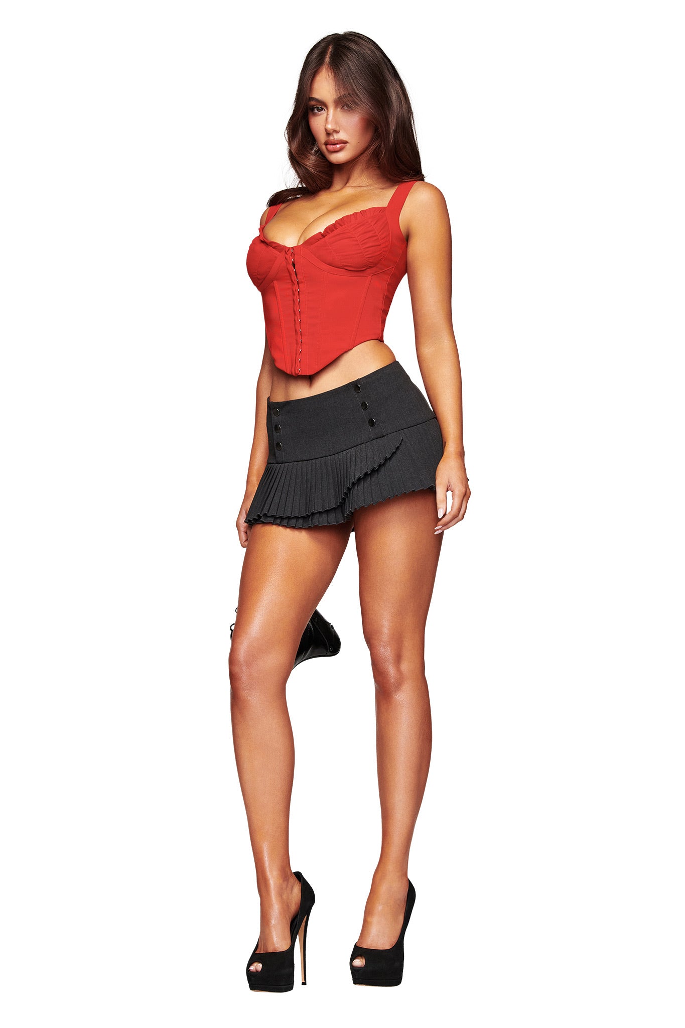A woman faces forward in the BRI CORSET - RICH RED by I.AM.GIA, styled with a short black pleated skirt and black high heels. Her long brown hair and confident expression stand out against a white background.