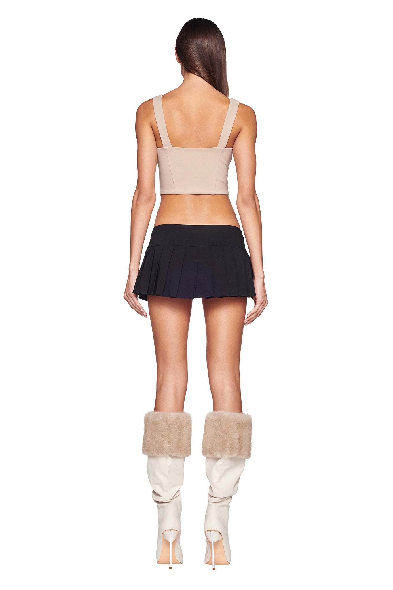 A woman stands facing away from the camera, wearing the I.AM.GIA BRI CORSET in TAN, paired with a black pleated mini skirt and knee-high white boots with beige fur trims. Her long, straight dark hair cascades down her back against a plain white background, emphasizing her chic I.AM.GIA-inspired ensemble.