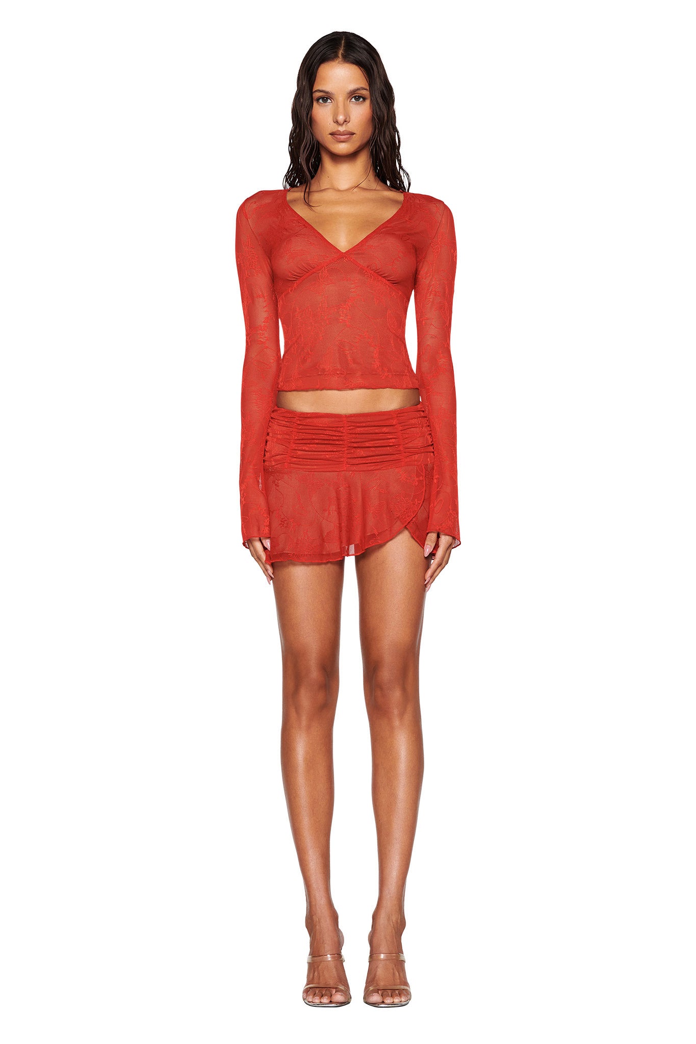 A person is standing against a white background wearing the I.AM.GIA YVETTE TOP in red paired with a matching red skirt featuring delicate butterfly lace detailing. They have long, wavy hair and are styled with transparent high-heeled shoes.