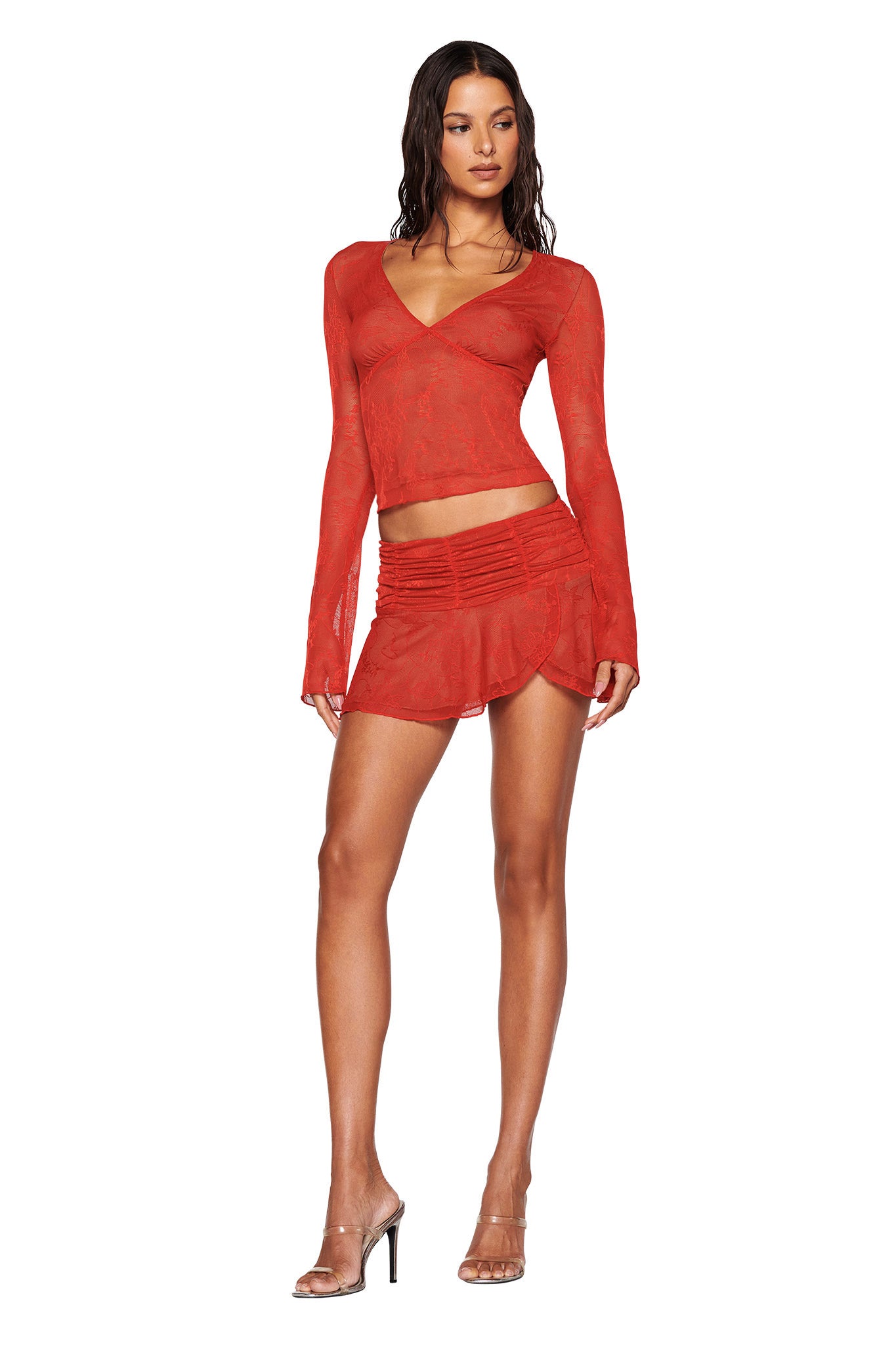 Wearing the YVETTE TOP in red from I.AM.GIA, a person pairs it with a matching mini skirt, both featuring intricate butterfly lace detailing. They stand against a white background in clear high-heeled sandals, their long wavy hair cascading down elegantly.