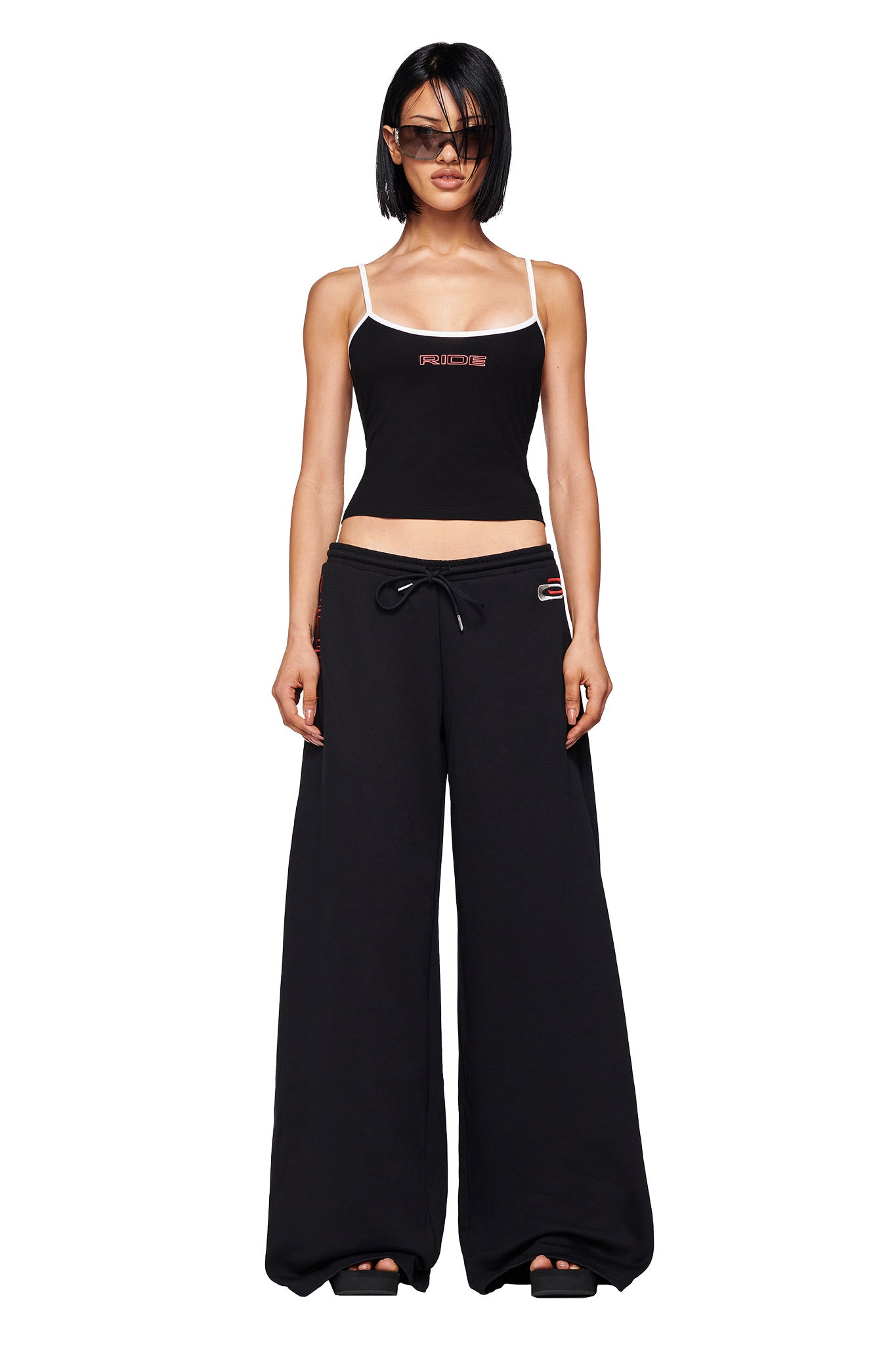 A woman in the I.AM.GIA SHAYLA TANK - BLACK with white straps, wide-leg black pants, black sandals, and sunglasses stands facing forward against a white background, featuring subtle I.AM.GIA graphic details.