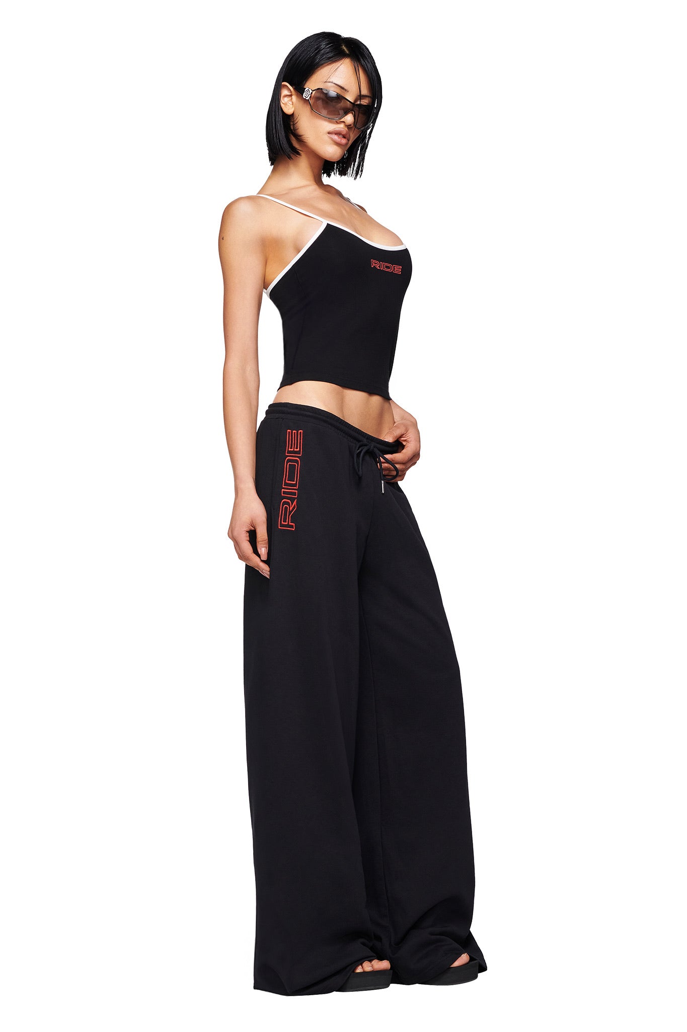 A woman stands confidently in I.AM.GIA's JUSTINE PANT - BLACK, featuring an ultra baggy fit, low-rise waist, and bold red "RIDE" placement print, paired with a black asymmetrical strap tank top and sunglasses against a white background.