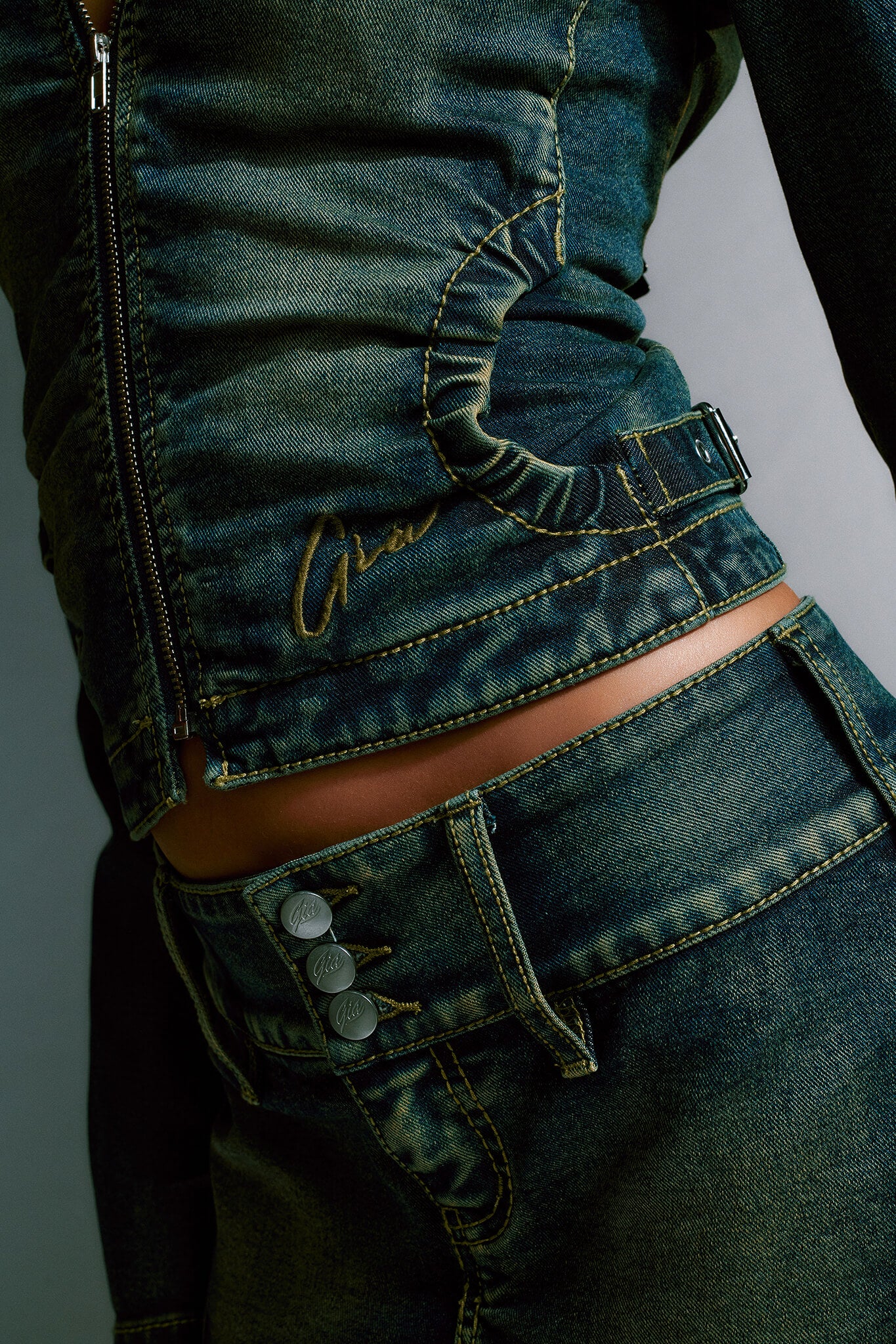 Close-up shot of a person wearing the NYKKO JACKET - BLUE and matching denim pants from I.AM.GIA. The jacket, in Denim Blue, features a zipper, visible seam design, and a metal buckle on the side. The pants are distinguished by three metal buttons on the left side, both garments showcasing detailed stitching.