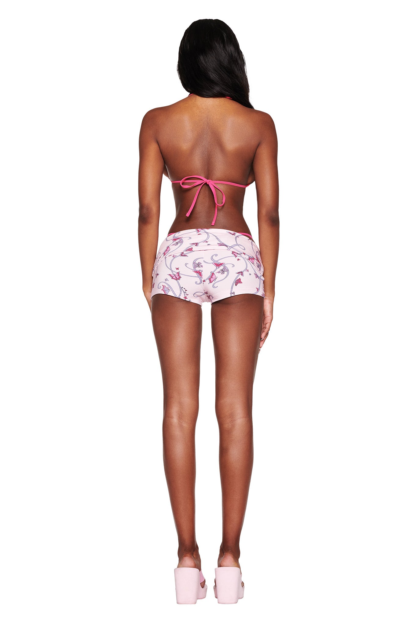 Rear view of a person with long hair wearing a pink halter bikini top and I.AM.GIA's RHIANNON SHORT in Butterfly Ribbon print, standing against a white background with pink platform sandals.
