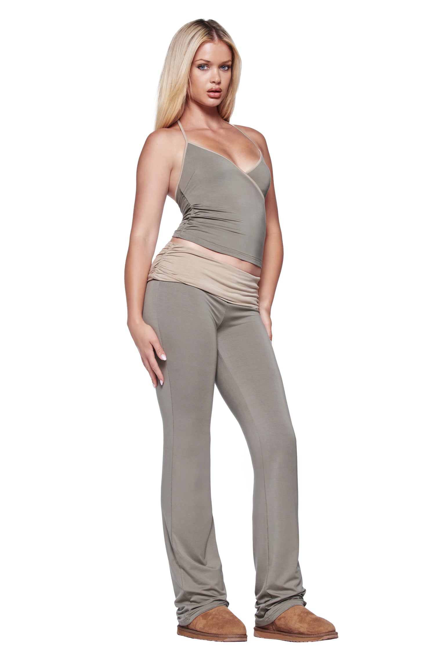 A woman with long blonde hair wears the I.AM.GIA JANNIS PANT in green, a taupe spaghetti strap top, a beige hip-accent belt, and brown shoes, standing forward against a white background.