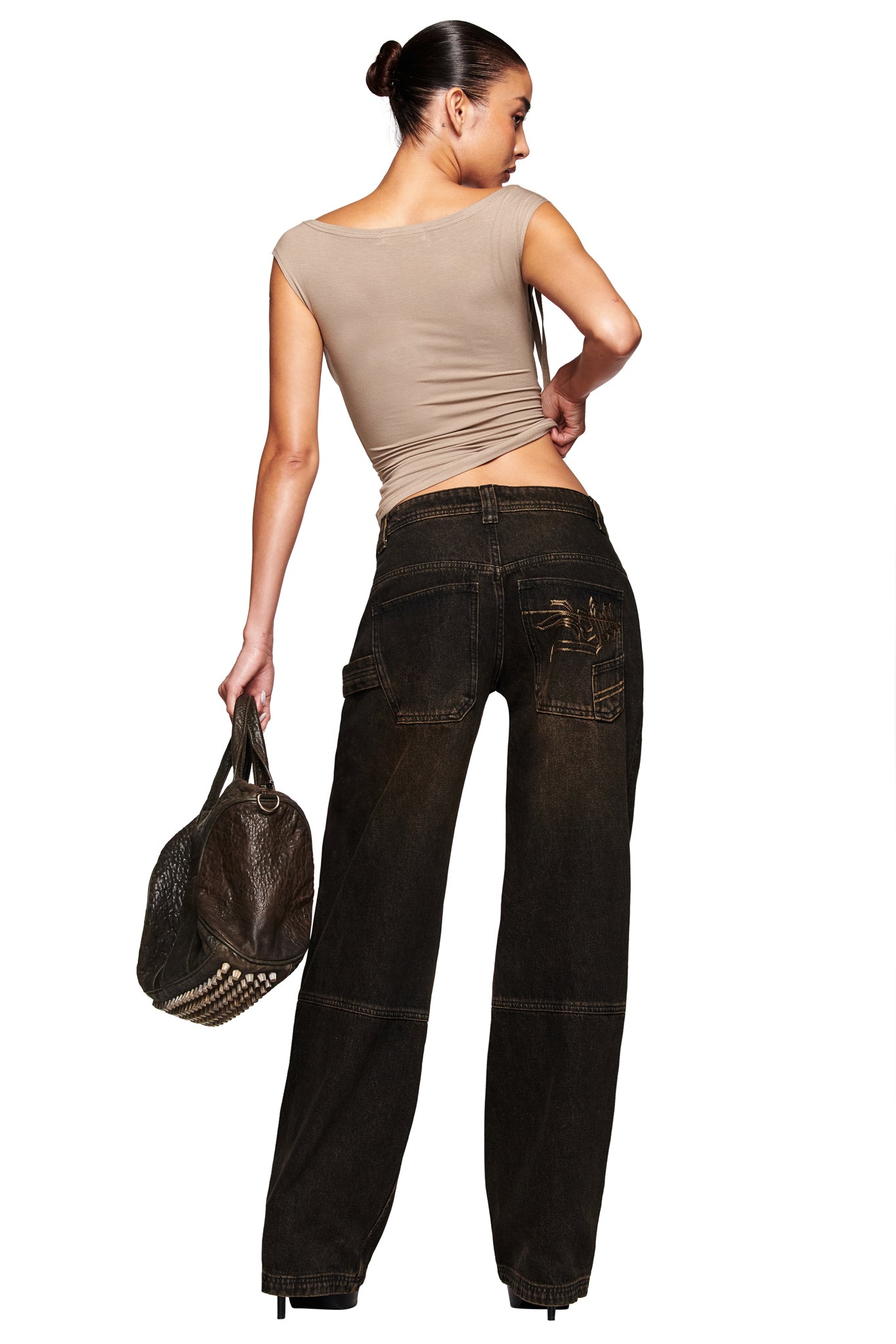 A woman with dark hair in a bun wears I.AM.GIA’s MISTY JEAN - BLACK, a beige cap-sleeve top, and holds a large brown textured handbag with metal studs, standing with her back to the camera.
